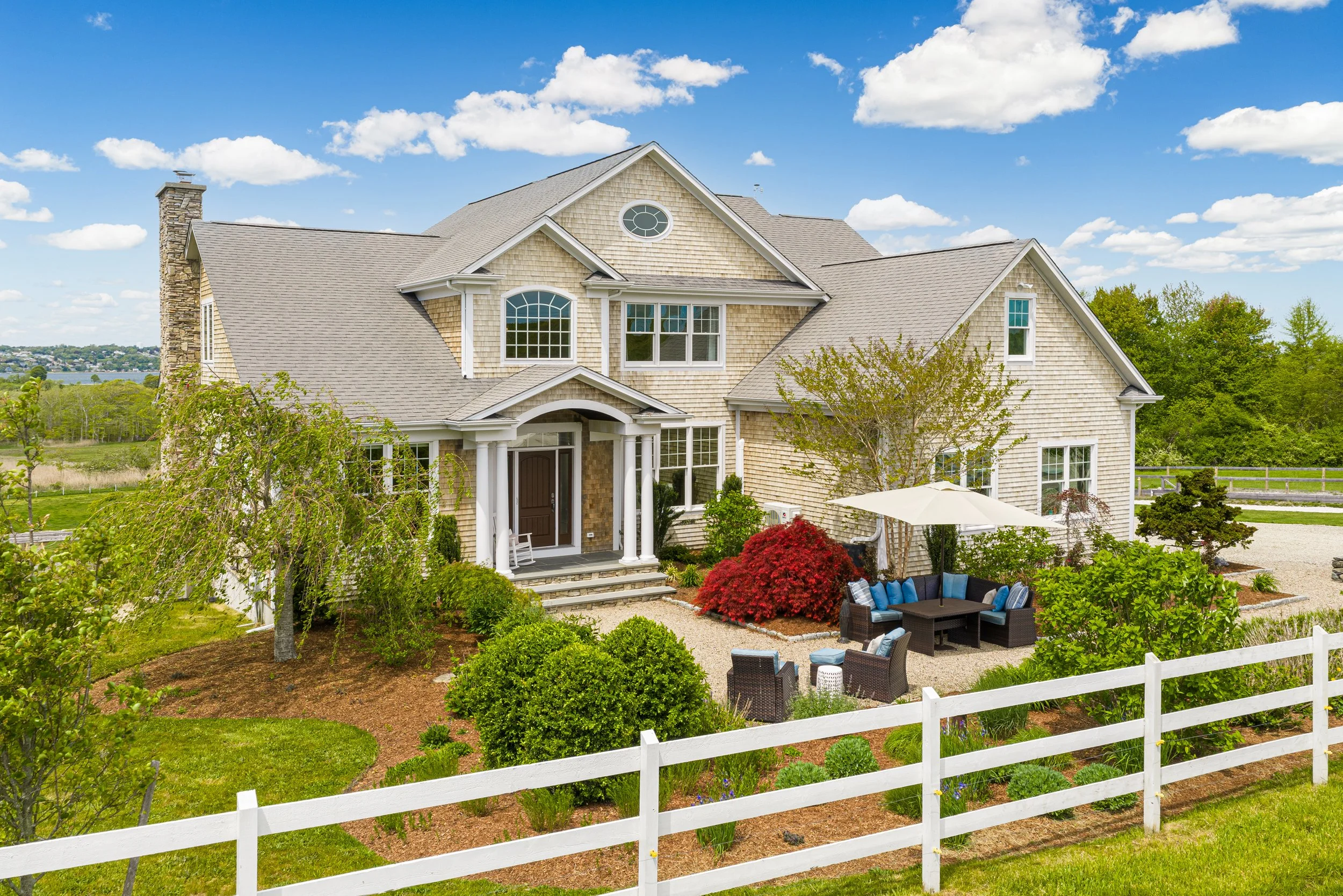 A large, multi-story house with traditional architectural style, surrounded by a landscaped garden with trees, shrubs, and outdoor seating, under a bright blue sky with scattered clouds.