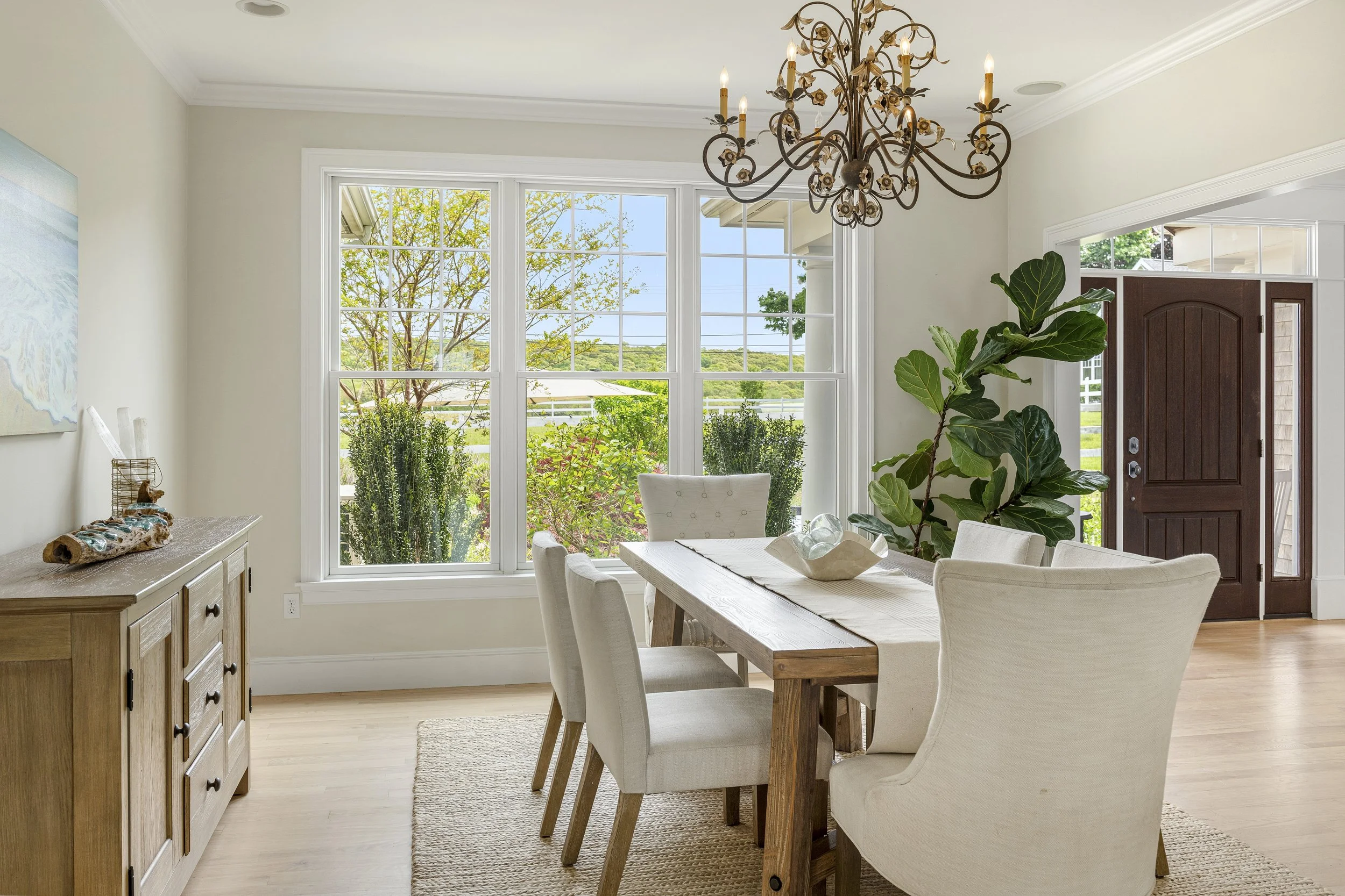 A dining area with a wooden table and six off-white upholstered chairs. Large windows with white trim let in natural light. A chandelier hangs from the ceiling. There is a large plant near the kitchen door. Outside, trees and a clear sky are visible.