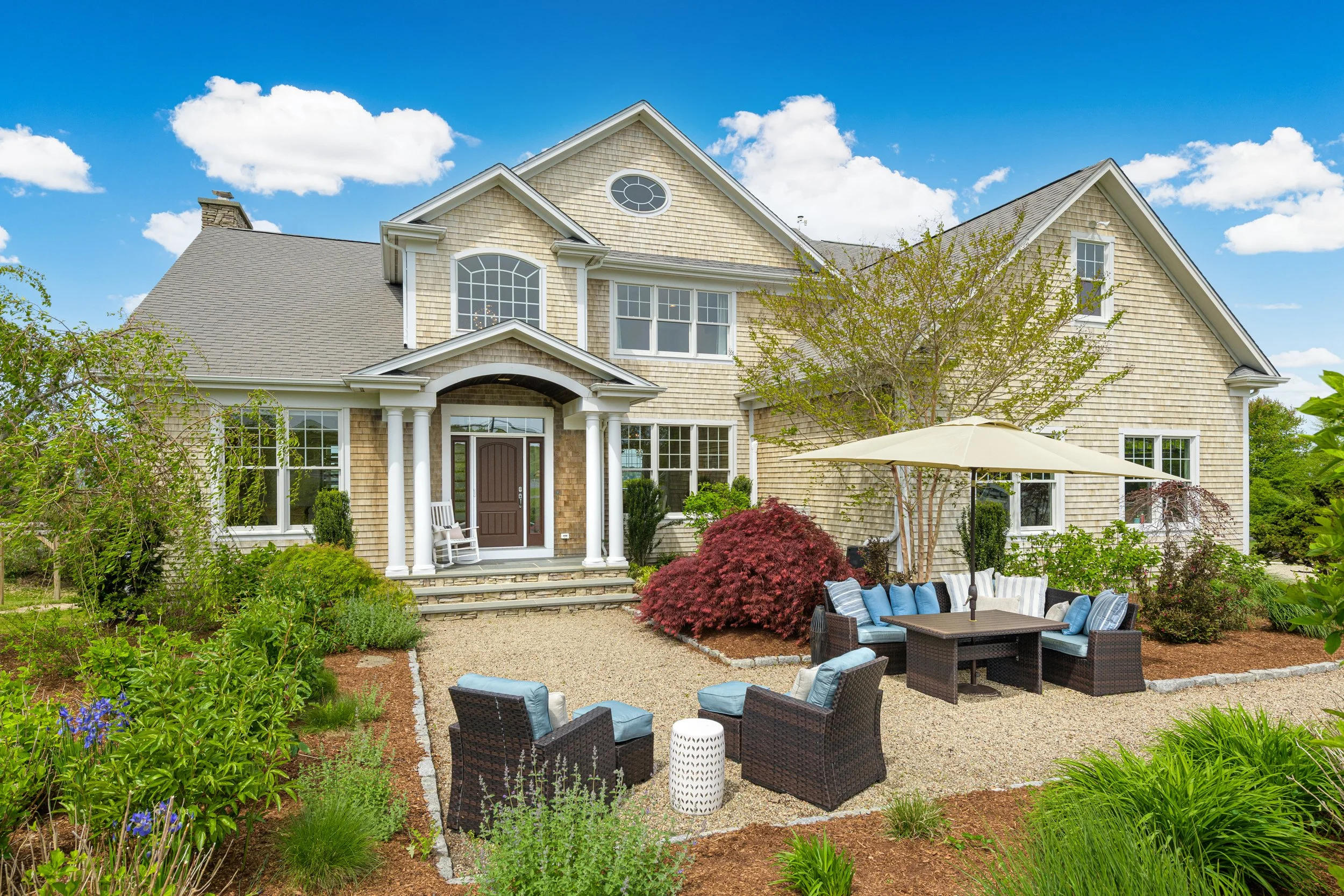A large beige house with a front porch surrounded by a lush landscaped garden with outdoor furniture and umbrellas, set under a blue sky with white clouds.