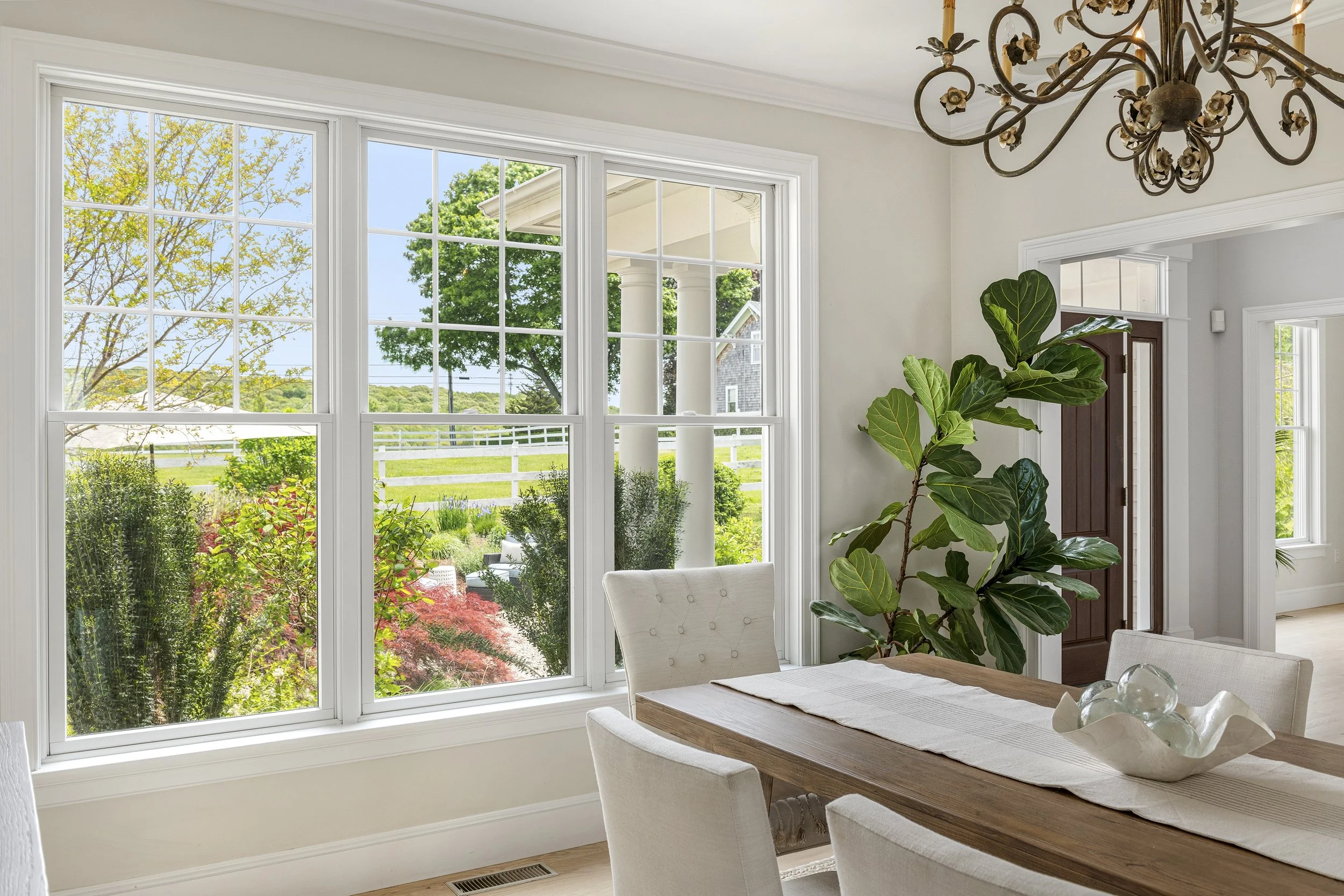 Bright dining area with large windows overlooking a garden with trees and shrubs, a white fence, and a blue sky. Inside, there are white upholstered chairs, a wooden dining table with a light-colored table runner, a decorative glass sculpture, a larg