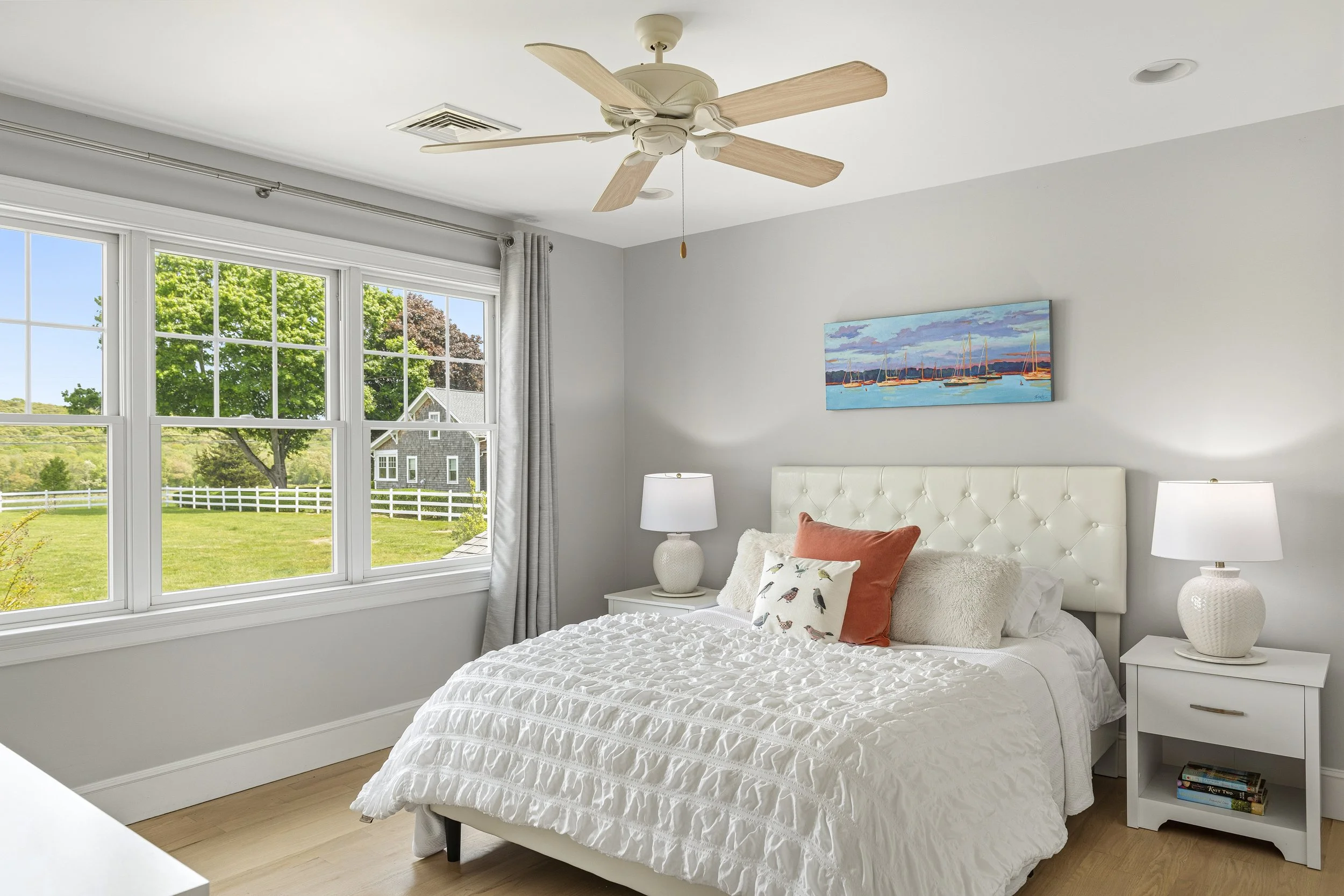 Bedroom with white bed, two white nightstands with lamps, large window showing green trees and a house outside, painting of sailboats above the bed, and a ceiling fan.