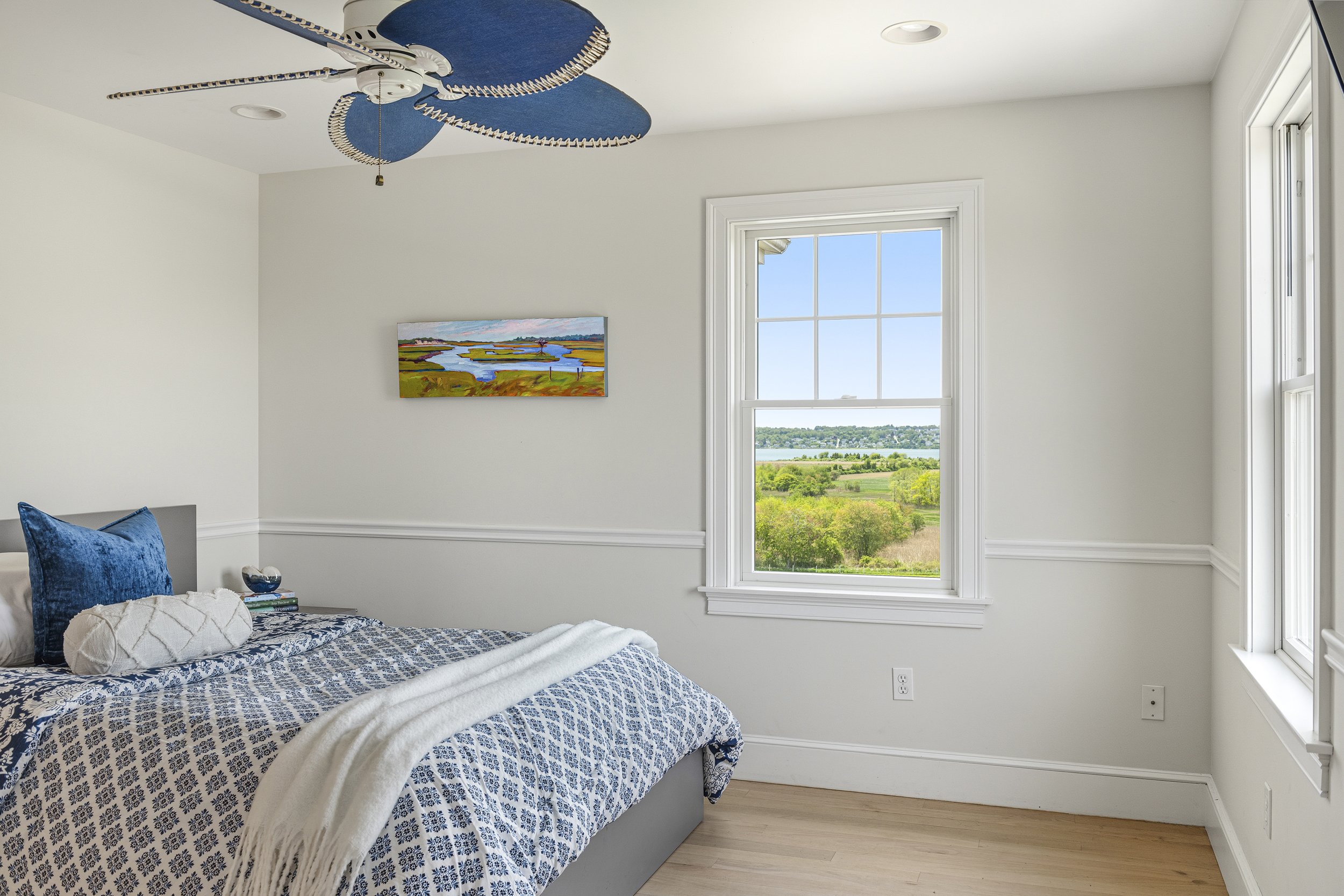 A bedroom with a bed covered in a blue and white patterned quilt, with pillows including a dark blue velvet one and a white textured one. There is a bedside table with books and a glass of water, and a large window with a view of green fields and tre