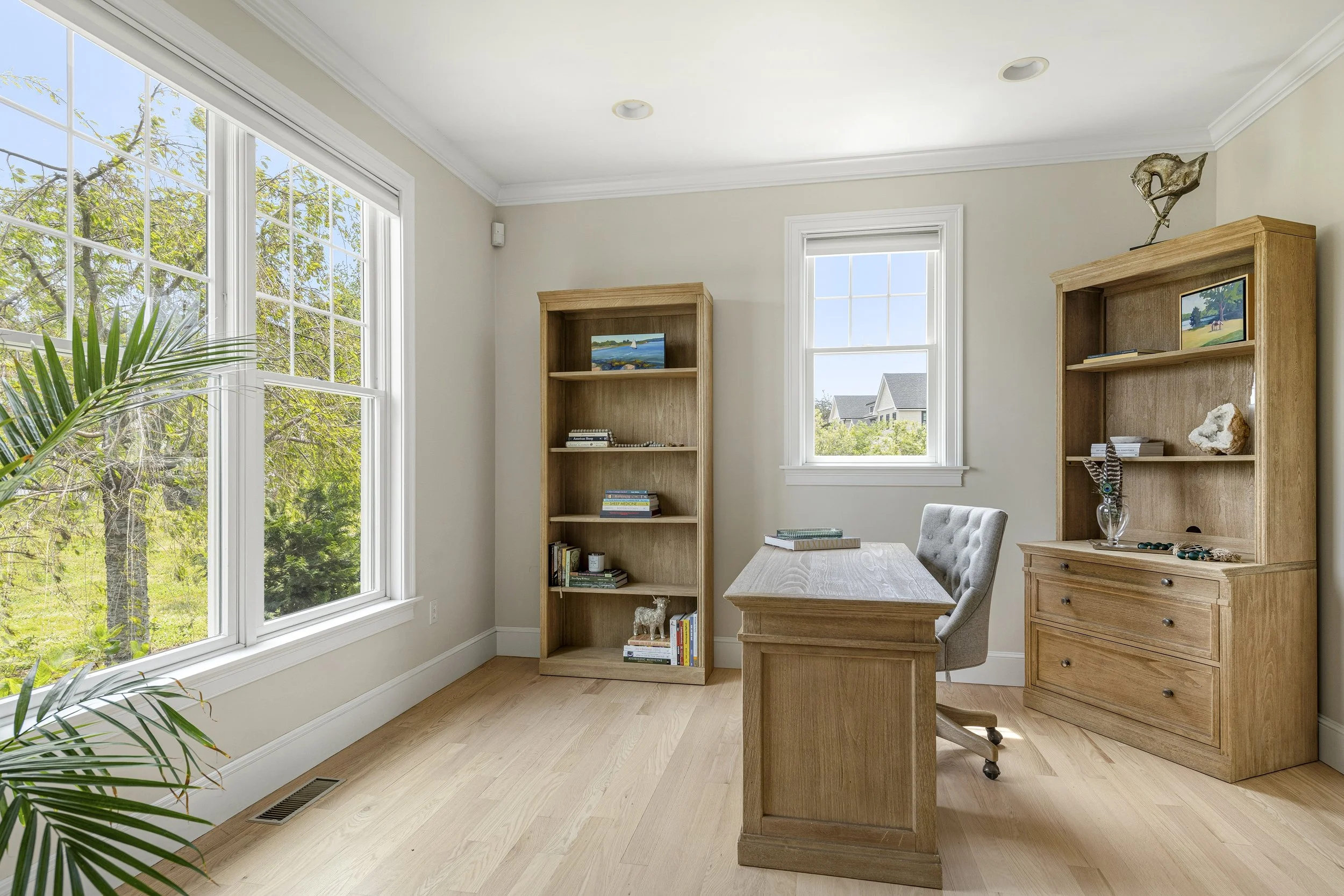 An office with large windows showing trees outside, a wooden desk with a cushioned office chair, two tall wooden bookshelves with books and decorative items, and hardwood floors.