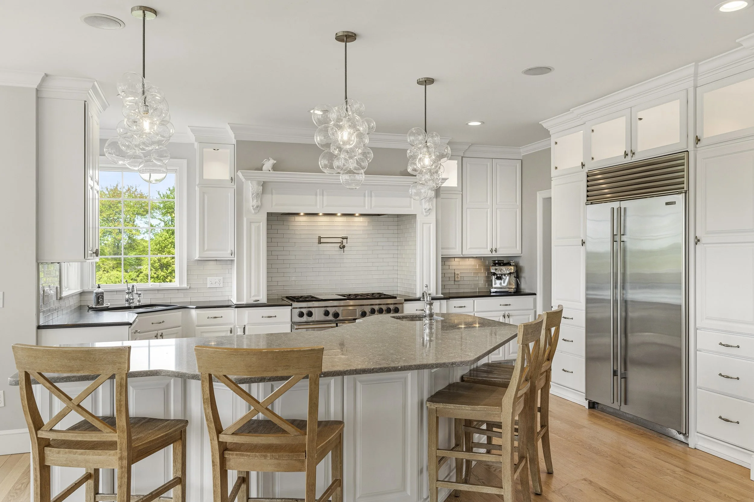 Bright kitchen with white cabinetry, a large window, a kitchen island with a granite countertop, and pendant lights made of glass bubbles hanging from the ceiling. There are wooden chairs around the island, a stainless steel refrigerator, and a coffe