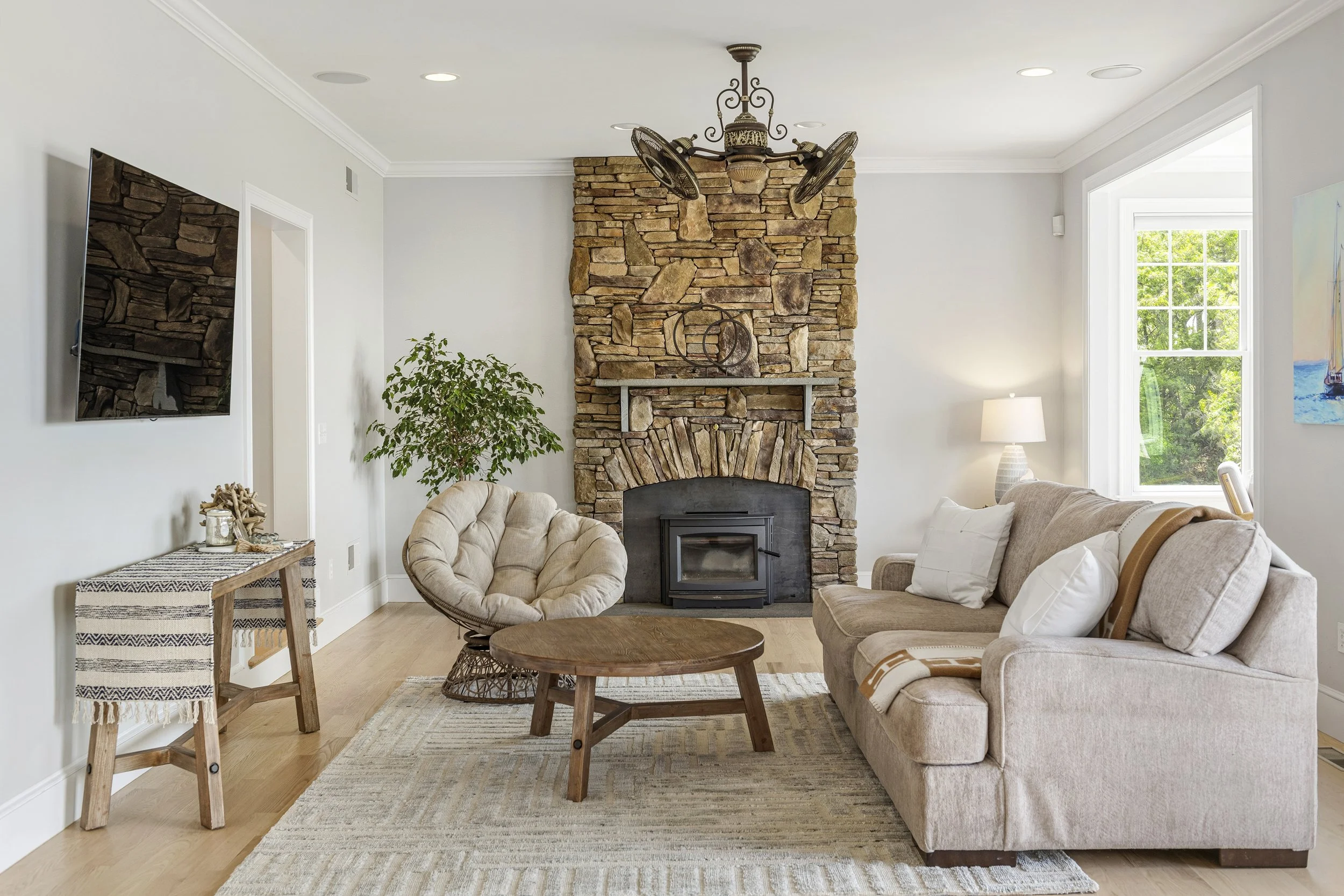 A cozy living room with a stone fireplace, a beige tufted armchair, a cream sofa with white pillows, a round wooden coffee table, and large windows with a view of green trees.