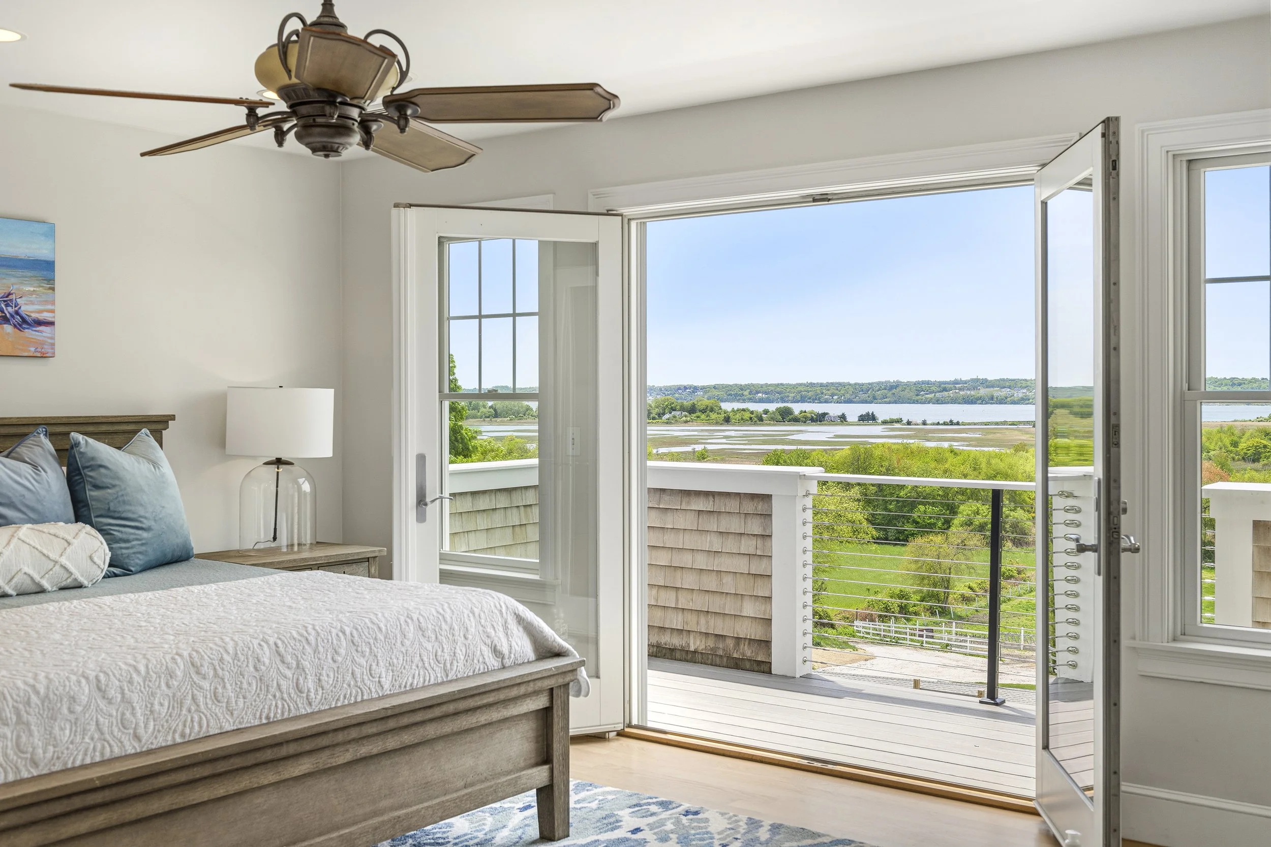 A bedroom with a bed, pillows, a nightstand with a lamp, and an open door leading to a balcony that overlooks a body of water and greenery.
