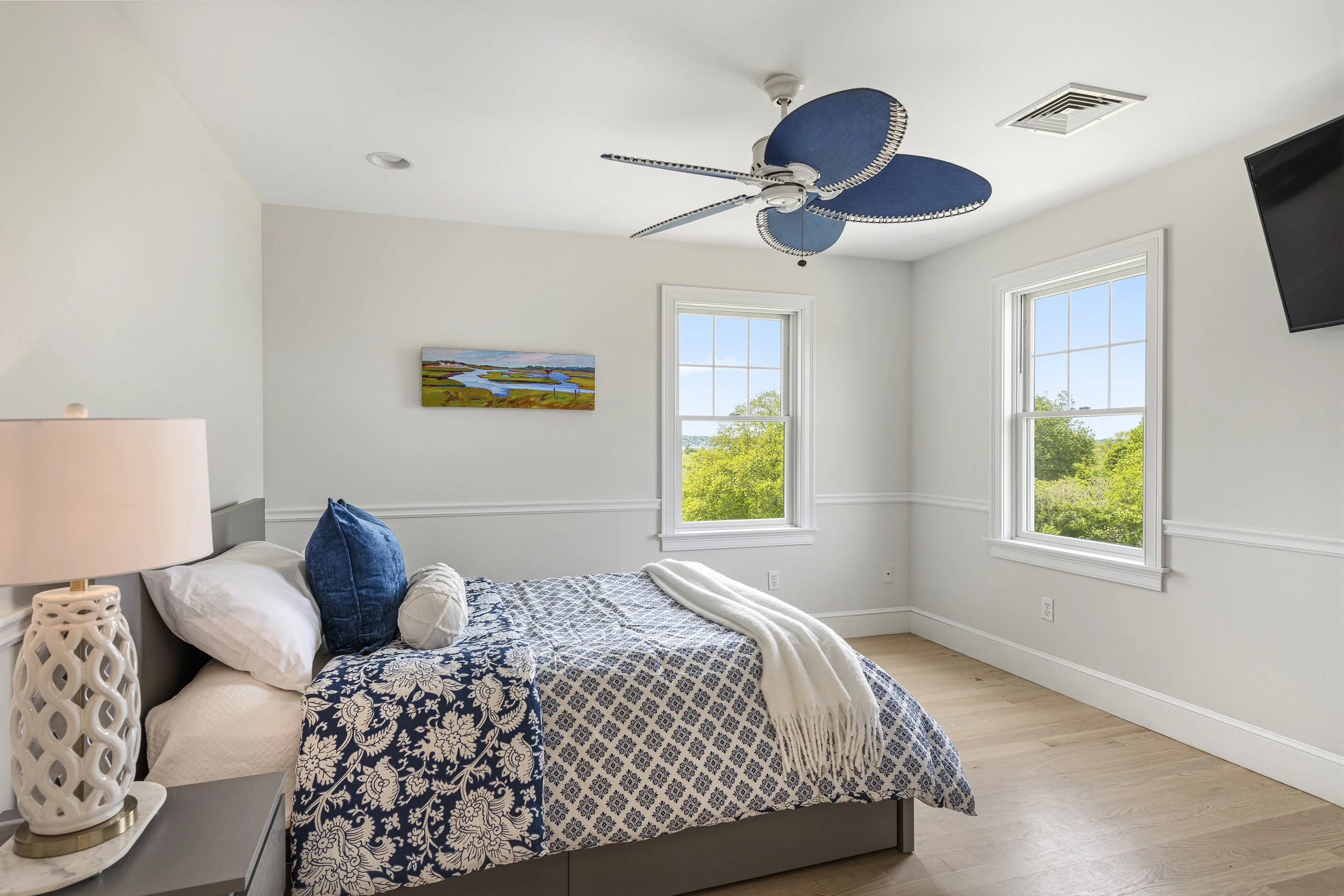 A bedroom with two large windows, a bed with blue and white patterned bedding, and a blue ceiling fan shaped like petals. There is a white table lamp with a lattice design on a nightstand next to the bed, and a small piece of colorful landscape artwo