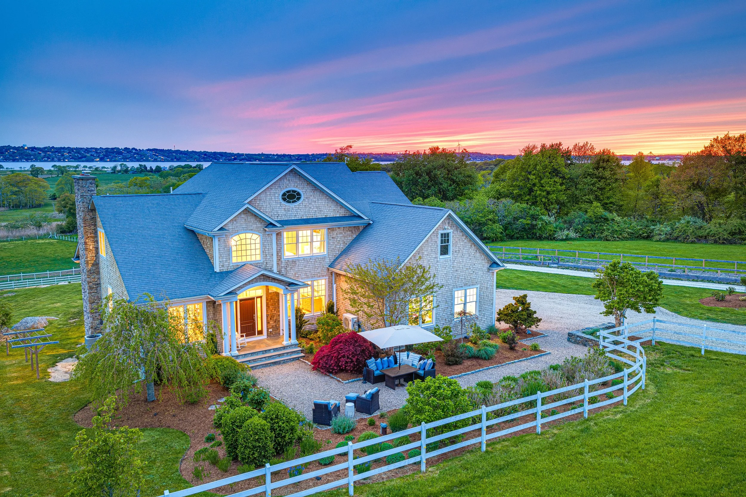 A two-story house with a gray roof, stone chimney, and illuminated windows, surrounded by a landscaped yard with outdoor furniture, trees, and a white fence. The sunset sky features pink and purple streaks over a distant body of water and hills.