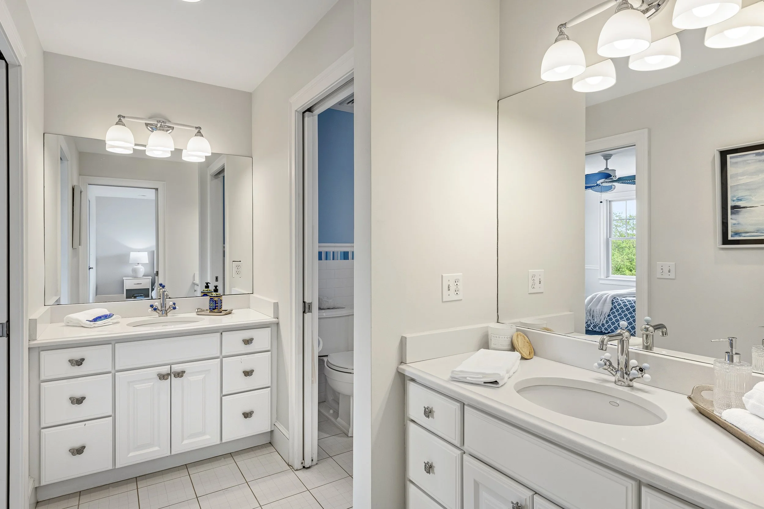 Bathroom with two white vanities, large mirror, and four-branched light fixture. One vanity has a sink with toiletries, and the other has a swimming pool drip tray with towels and a soap dish. To the side, a window and a partially visible shower/tub 