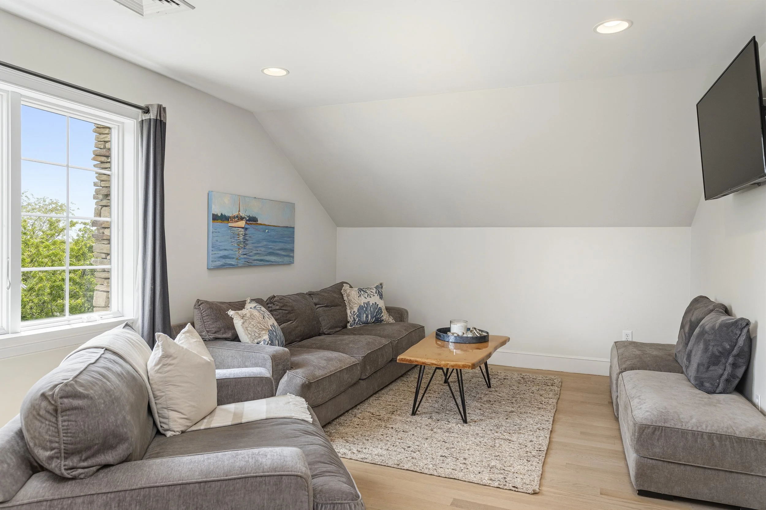Living room with large windows, gray sofas, decorative pillows, a wooden coffee table, a colorful nautical painting, and a wall-mounted television.
