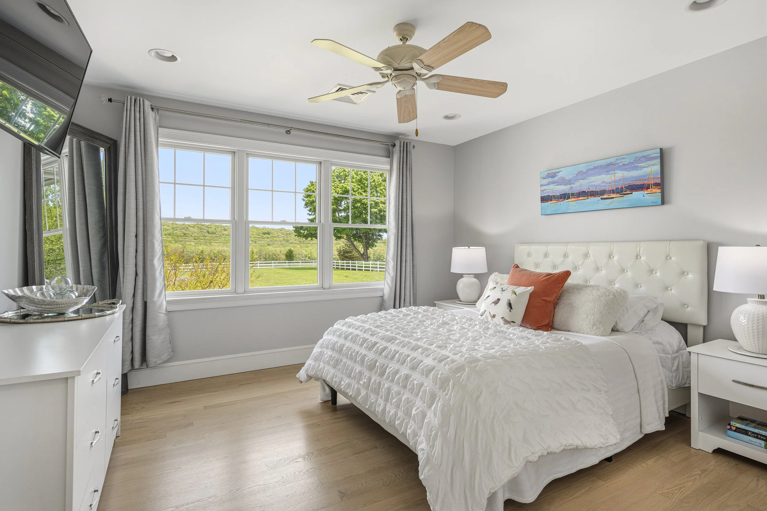 A bright bedroom with large window overlooking green landscape, white bed with textured bedding, decorative pillows, white nightstand, lamps, wall art of sailboats, ceiling fan, and dresser with glass bowl and mirror.
