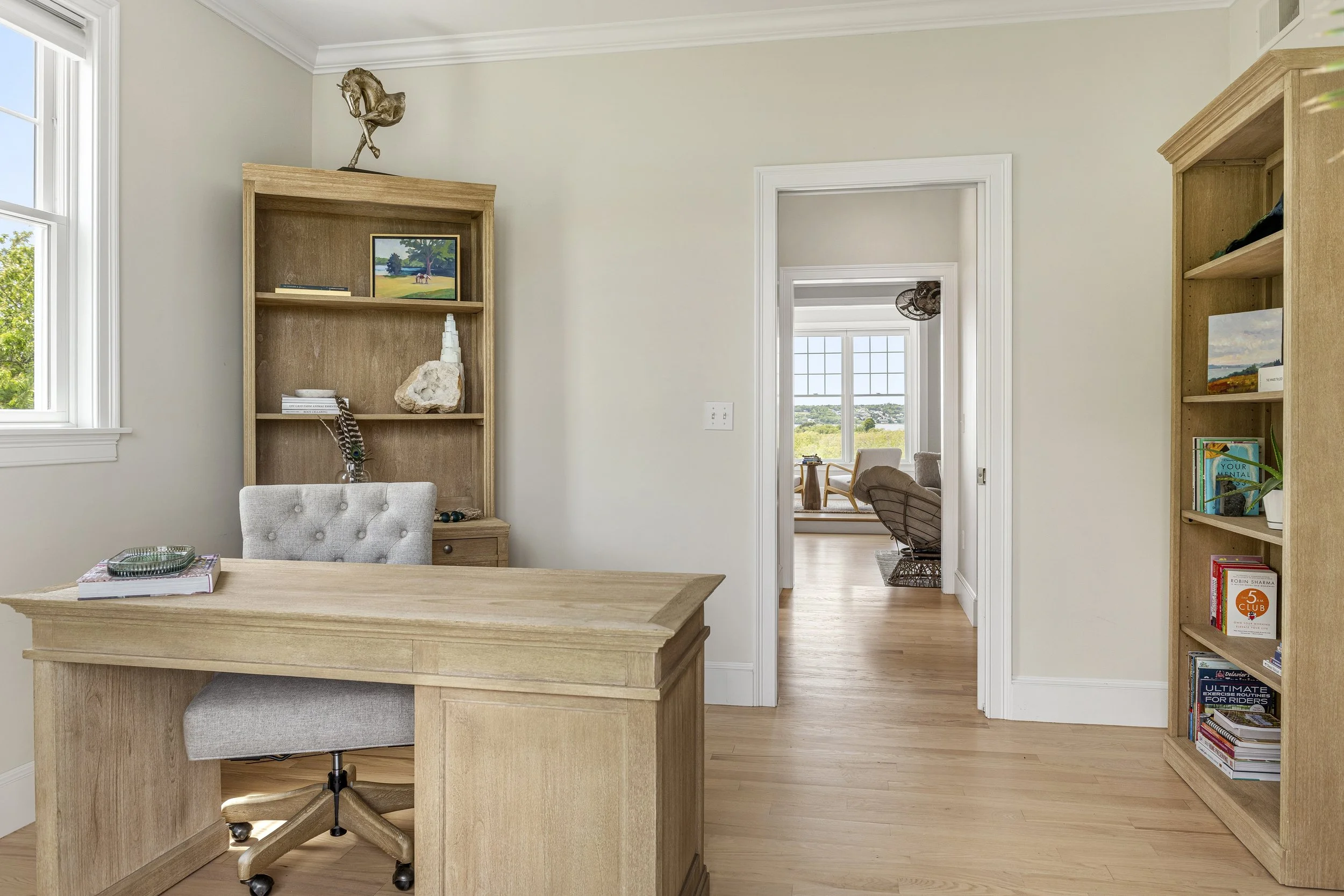 A home interior with a wooden desk and a gray upholstered office chair. There are bookshelves with books and decorative items, and an open doorway leading to a room with large windows showing a landscape view outside.