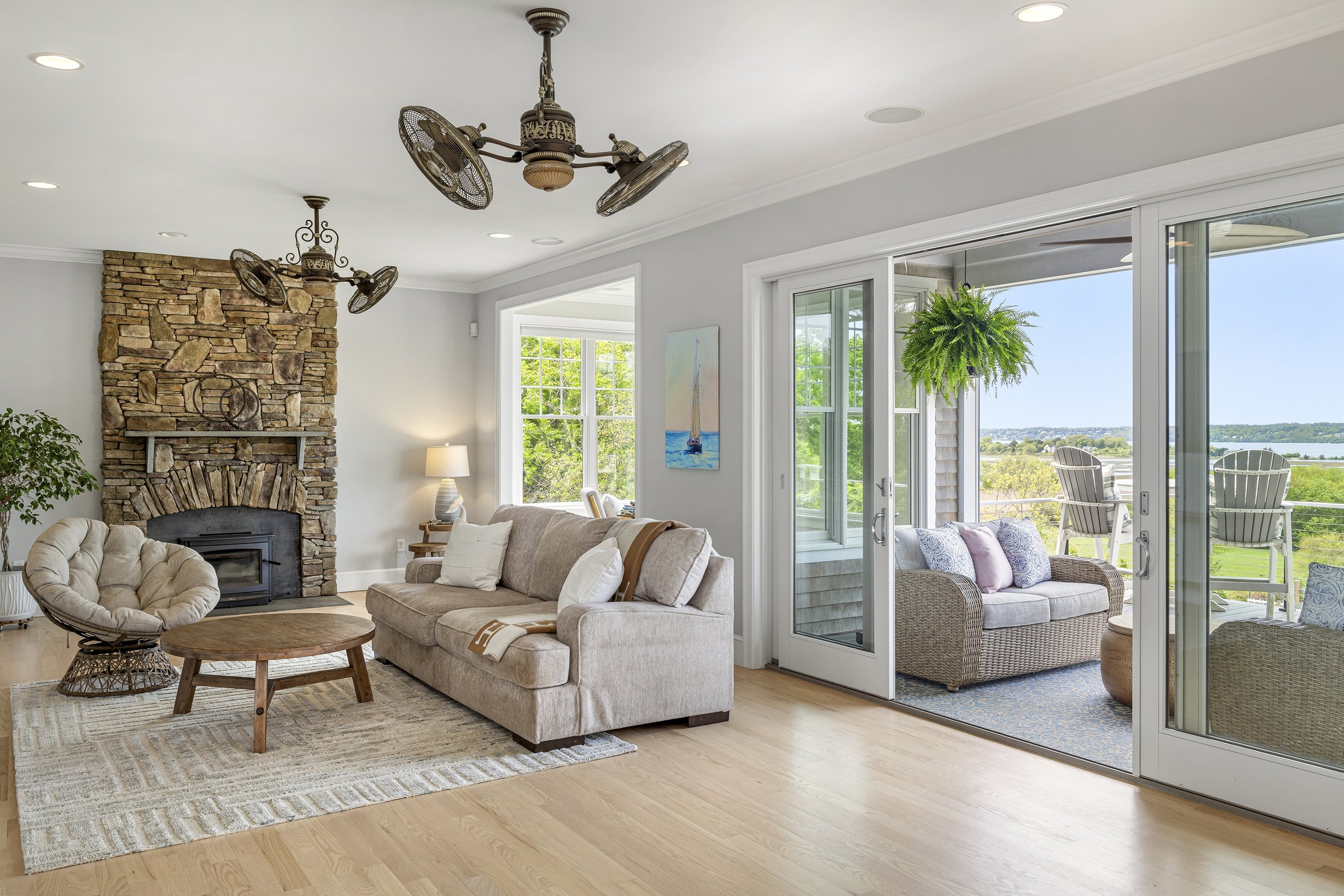 Living room with beige couches, a stone fireplace, large sliding glass door leading to a balcony with outdoor seating and a view of a river and green landscape.