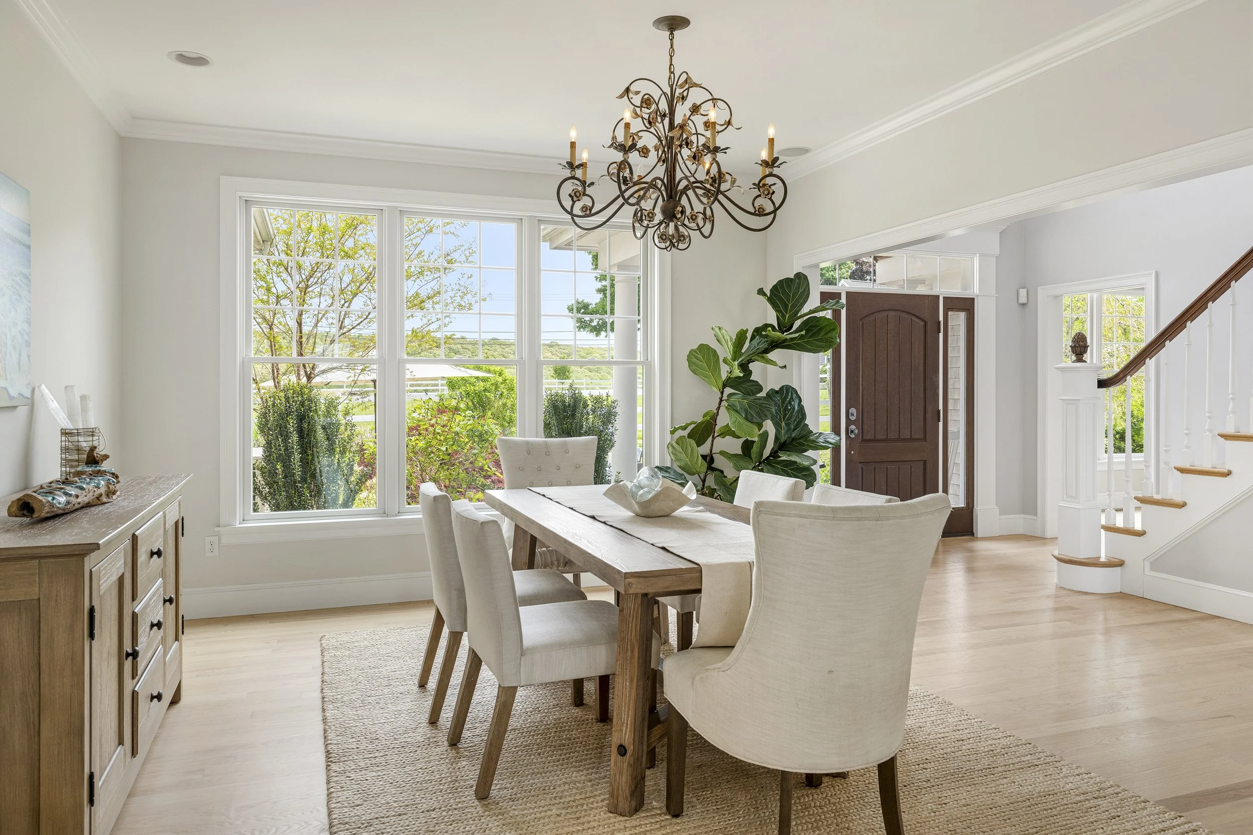 Bright dining room with large windows showing a green outdoor landscape, featuring a wooden dining table surrounded by cream-colored chairs, a chandelier hanging from the ceiling, and a sideboard with decorative items