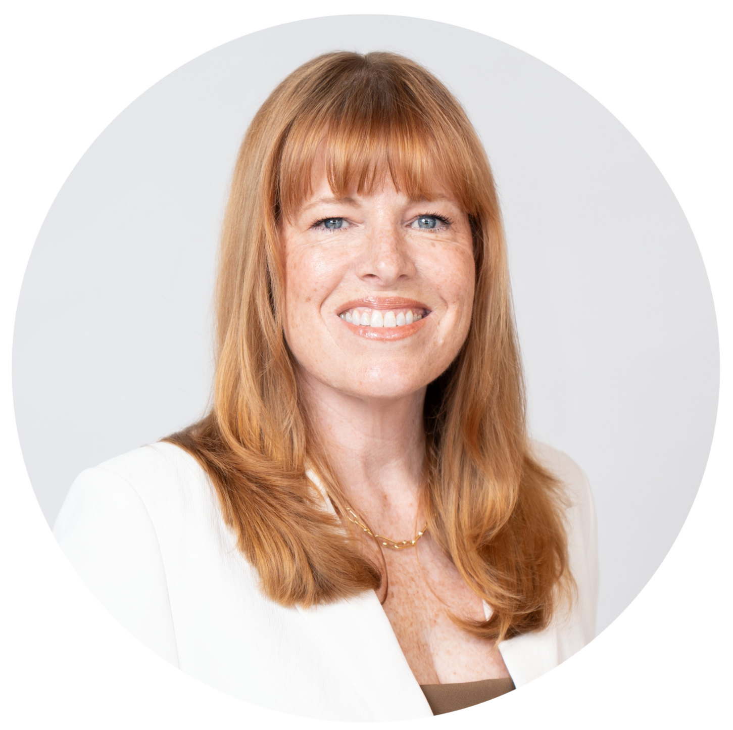 Portrait of a smiling woman with red hair, blue eyes, wearing a white blazer and a gold necklace, against a plain light background.