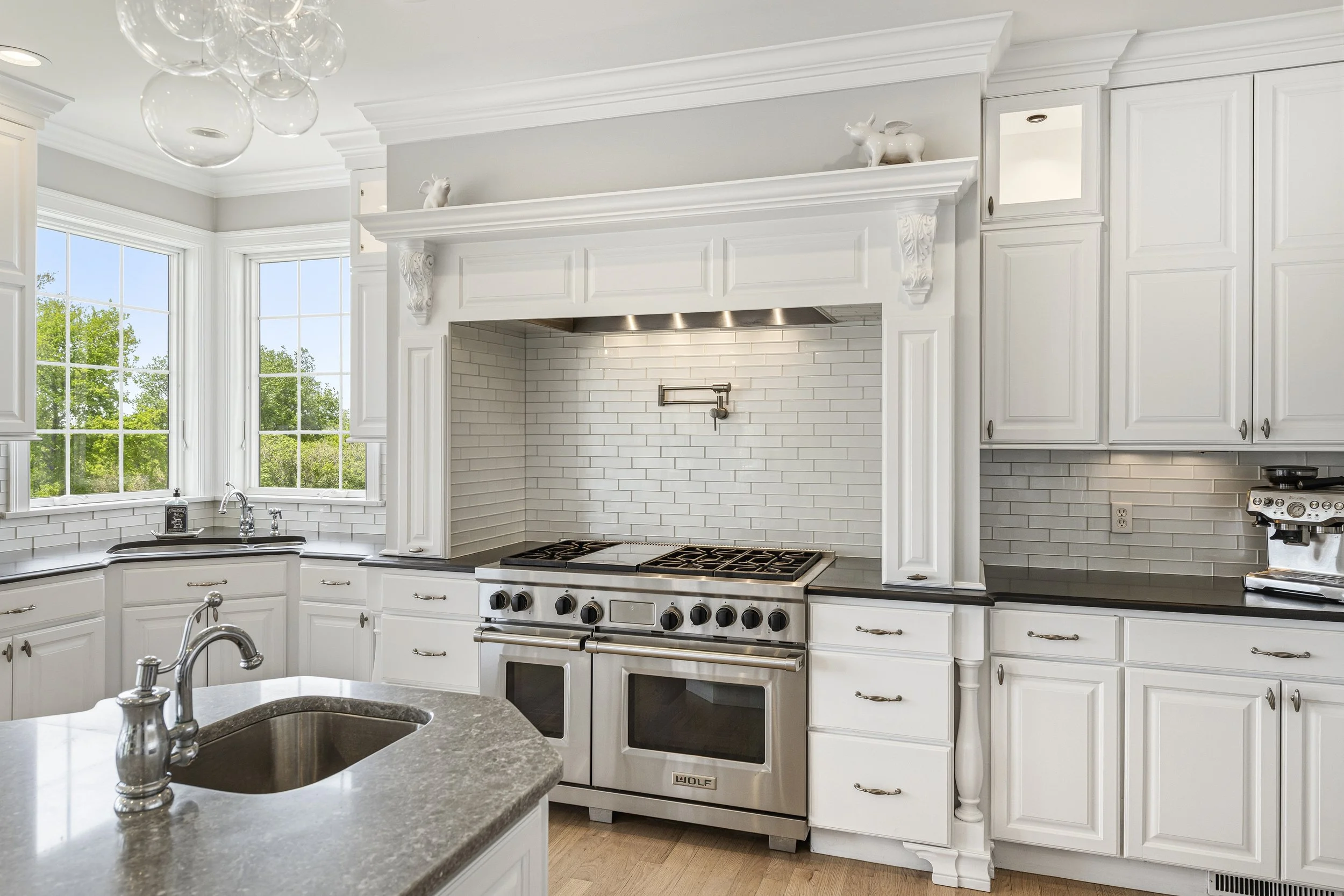 White kitchen with large windows, white cabinetry, a stove with a vent hood, a gray countertop, and a sink with a chrome faucet.