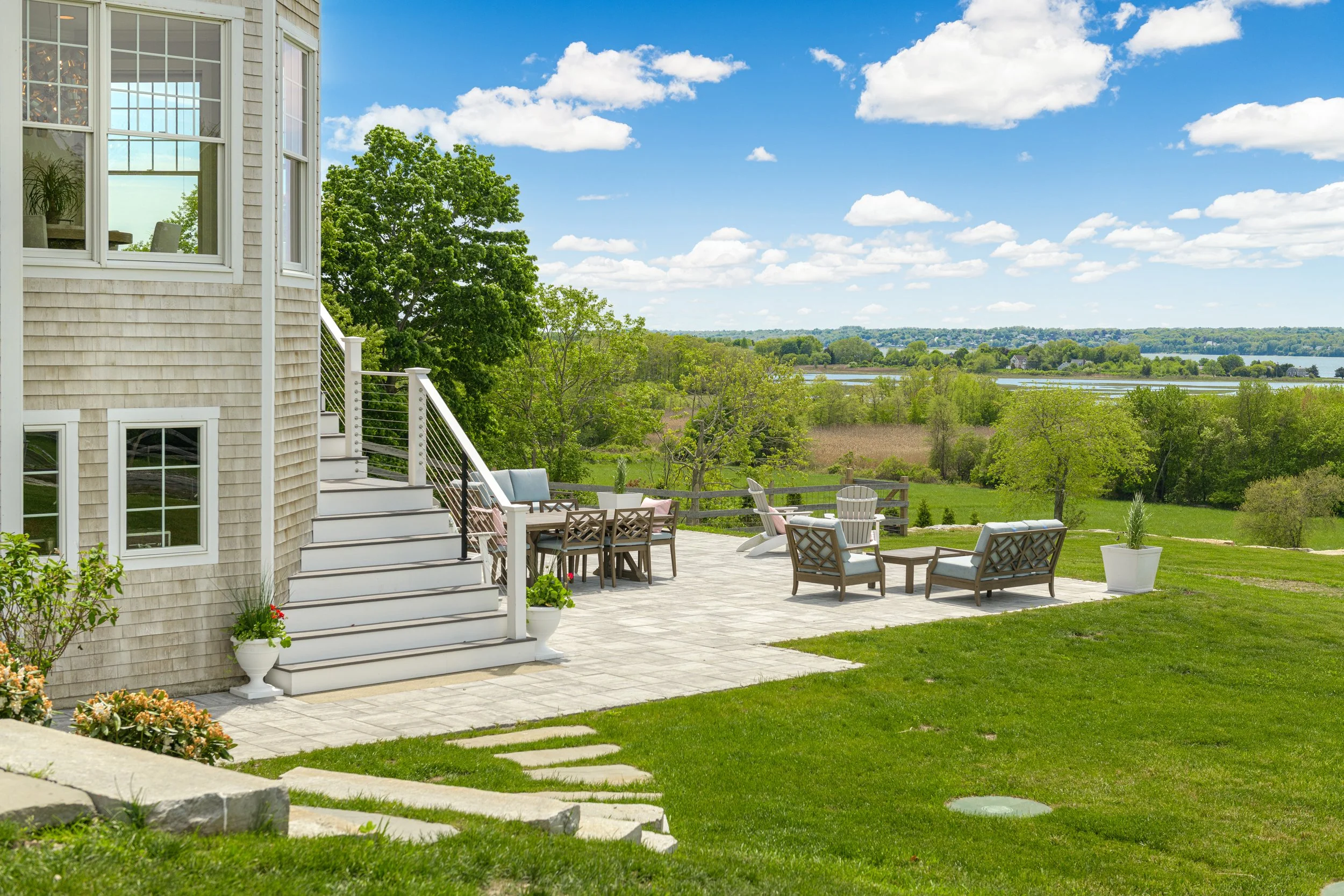 A backyard patio area with outdoor furniture, overlooking a scenic view of trees, a lake, and a partly cloudy sky.
