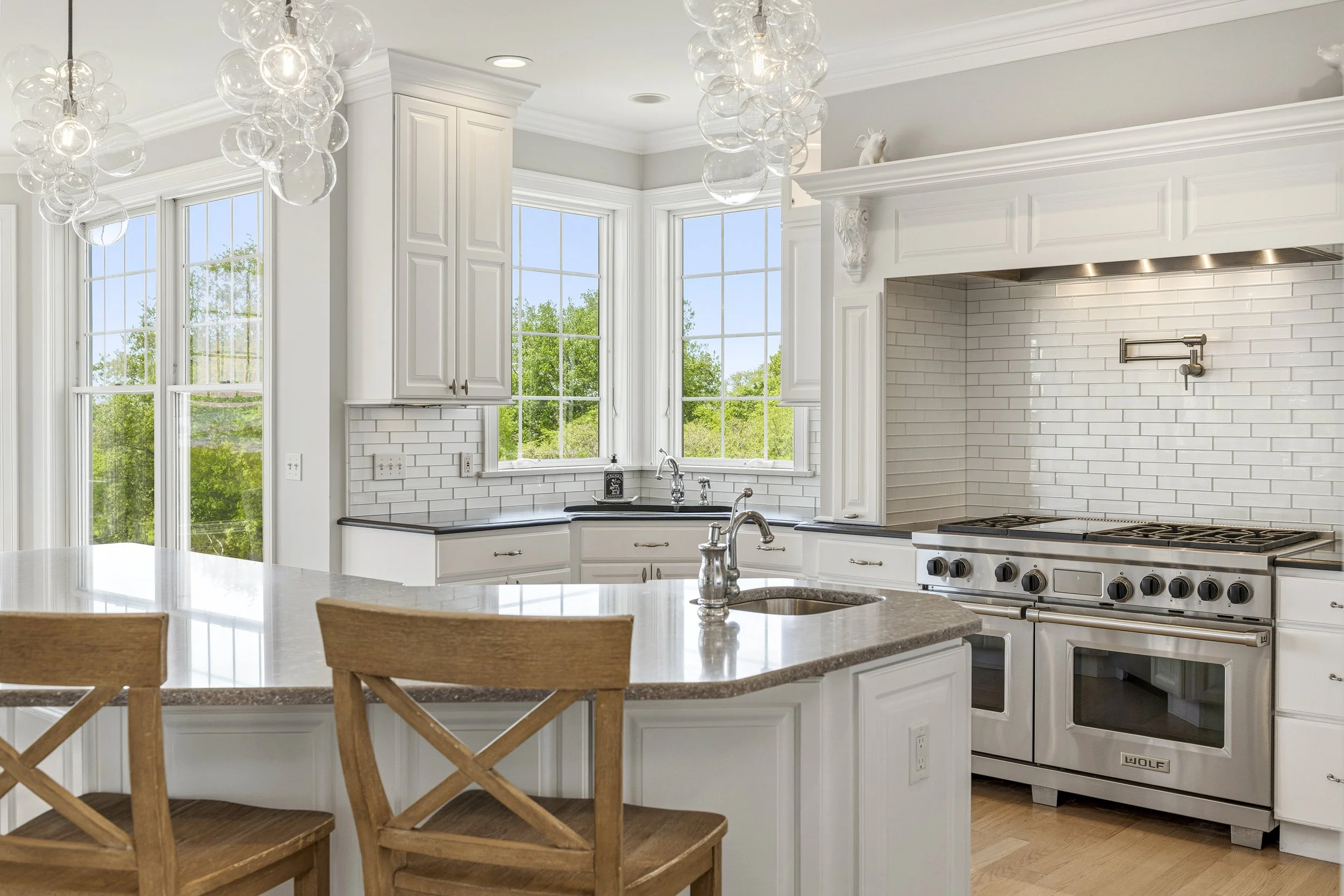 Modern kitchen with white cabinets, subway tile backsplash, large windows with scenic views, and a stainless steel stove. A light-colored island with a granite top and wooden chairs are in the foreground.