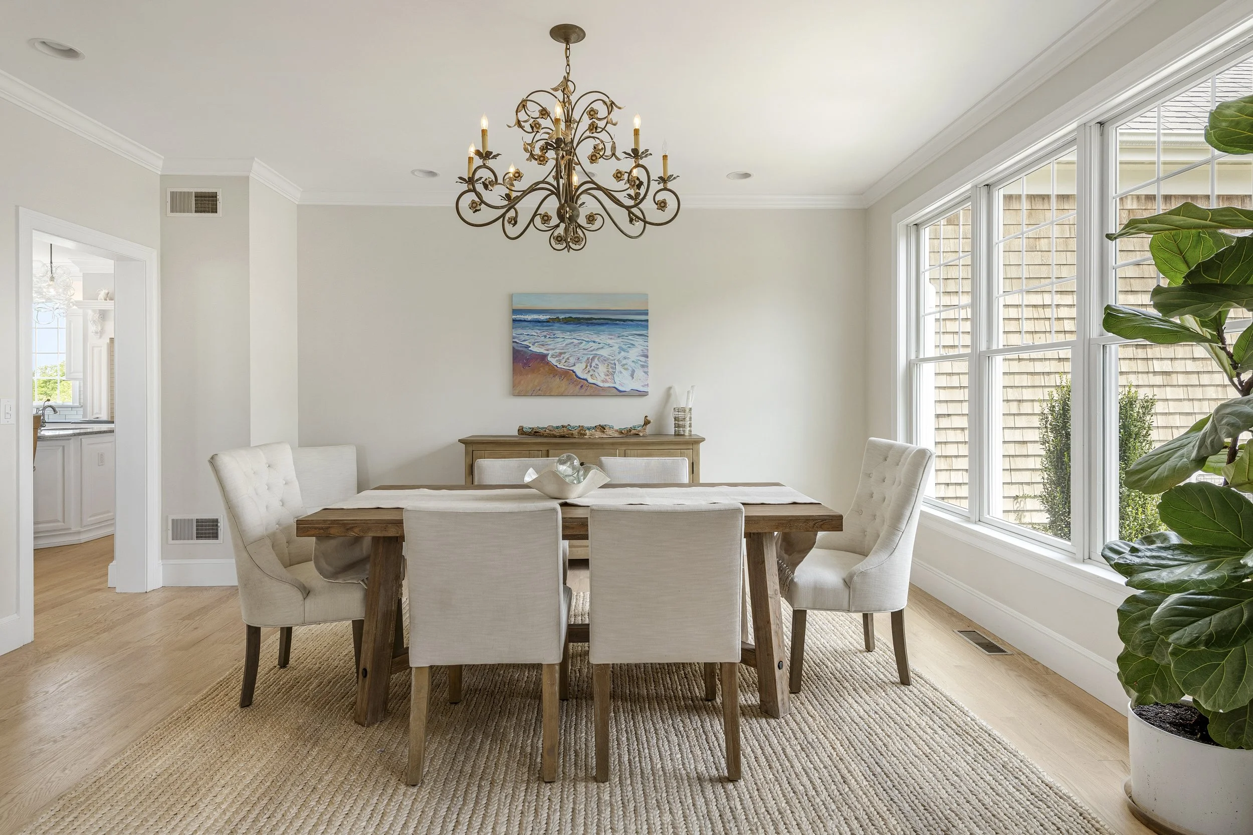 A dining room with a large window, a wooden table with a white table runner, eight cream upholstered chairs, a chandelier, a sideboard with a painting of the ocean, a sculpture, and a large green plant.