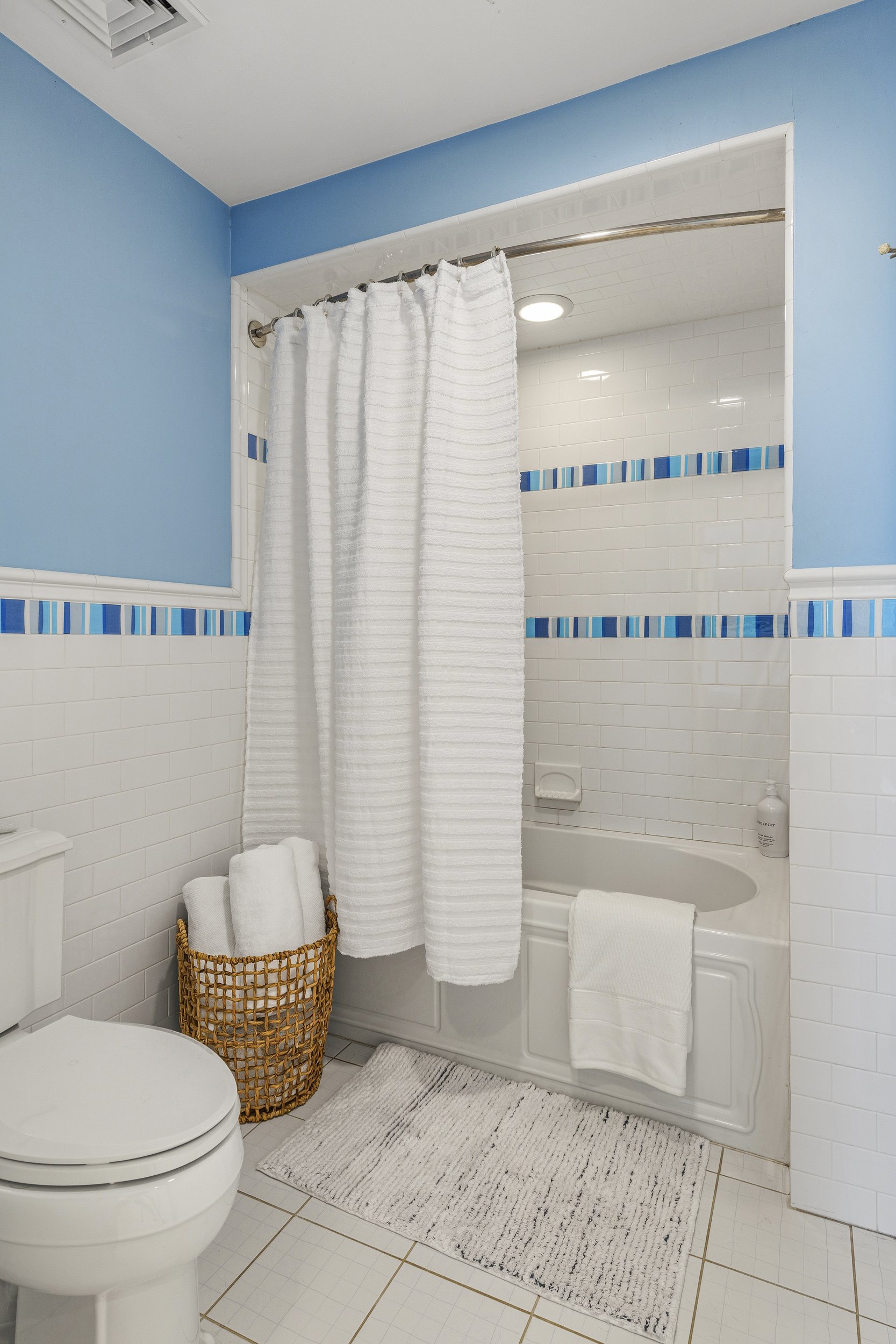 A bathroom with white tiled walls accented by a horizontal row of blue mosaic tiles. There is a white bathtub with a curved front, partially covered by a white towel hanging over the edge. A white shower curtain on a curved rod partially covers the b