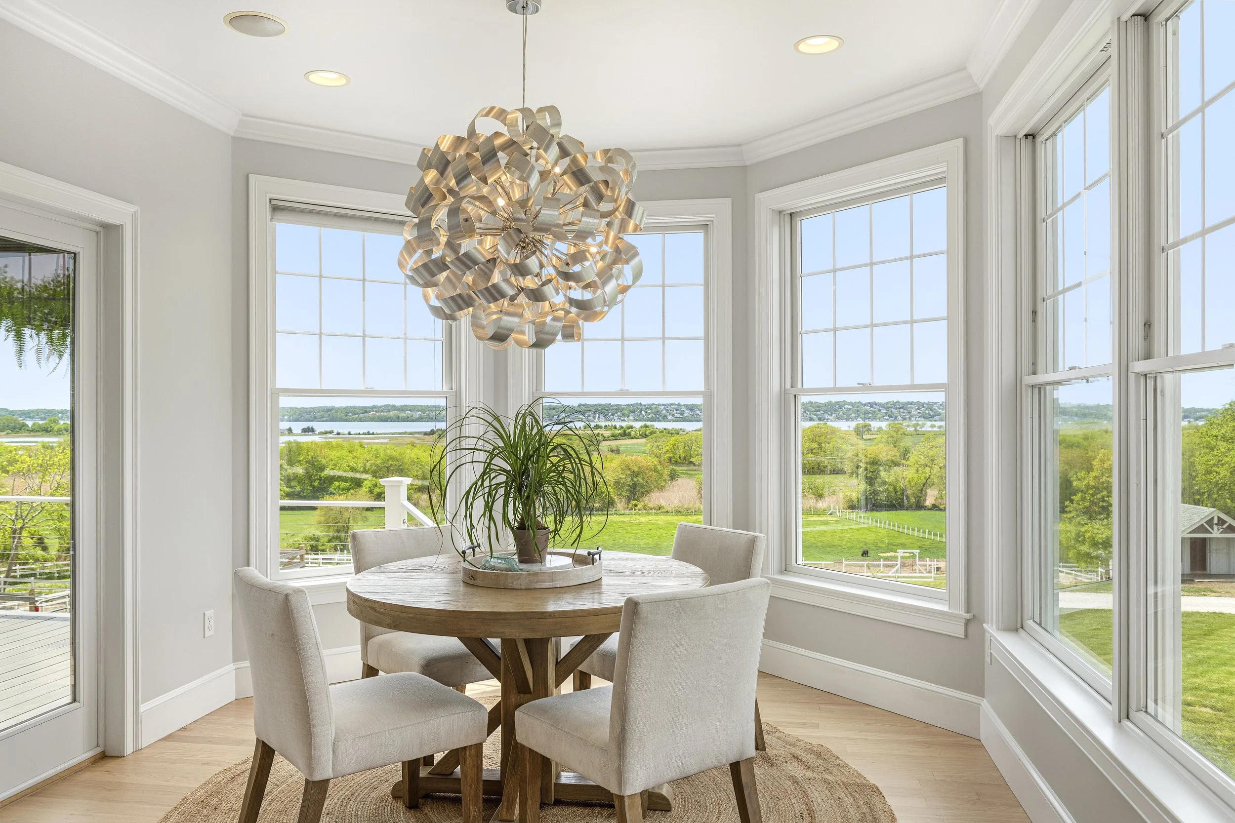 A dining area with a round wooden table, four beige upholstered chairs, a potted plant, and large windows overlooking a green landscape with water and trees.