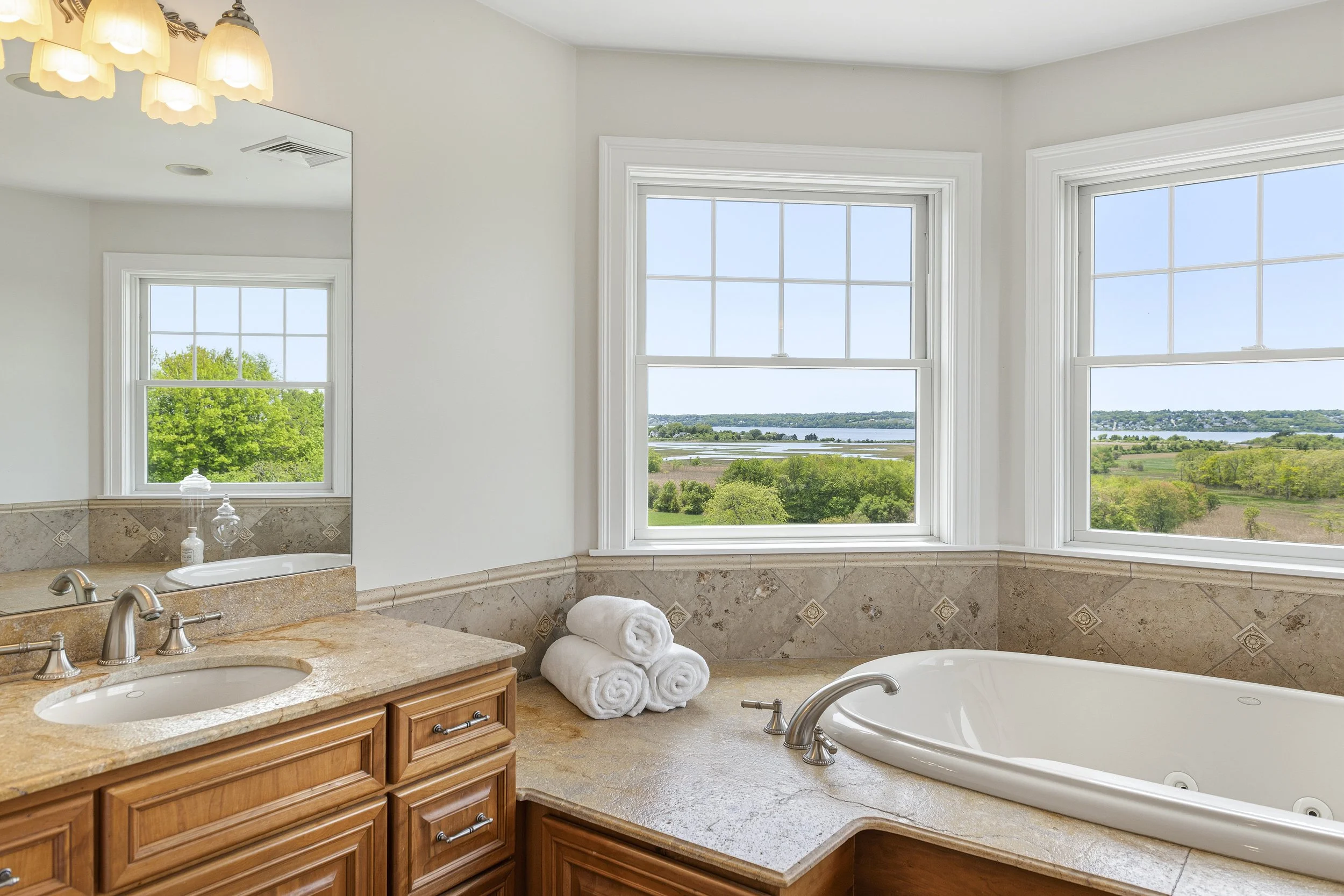 A bathroom with a view of green trees and a lake through three large windows. The bathroom features a stone countertop with sinks, a mirror, and rolled white towels.