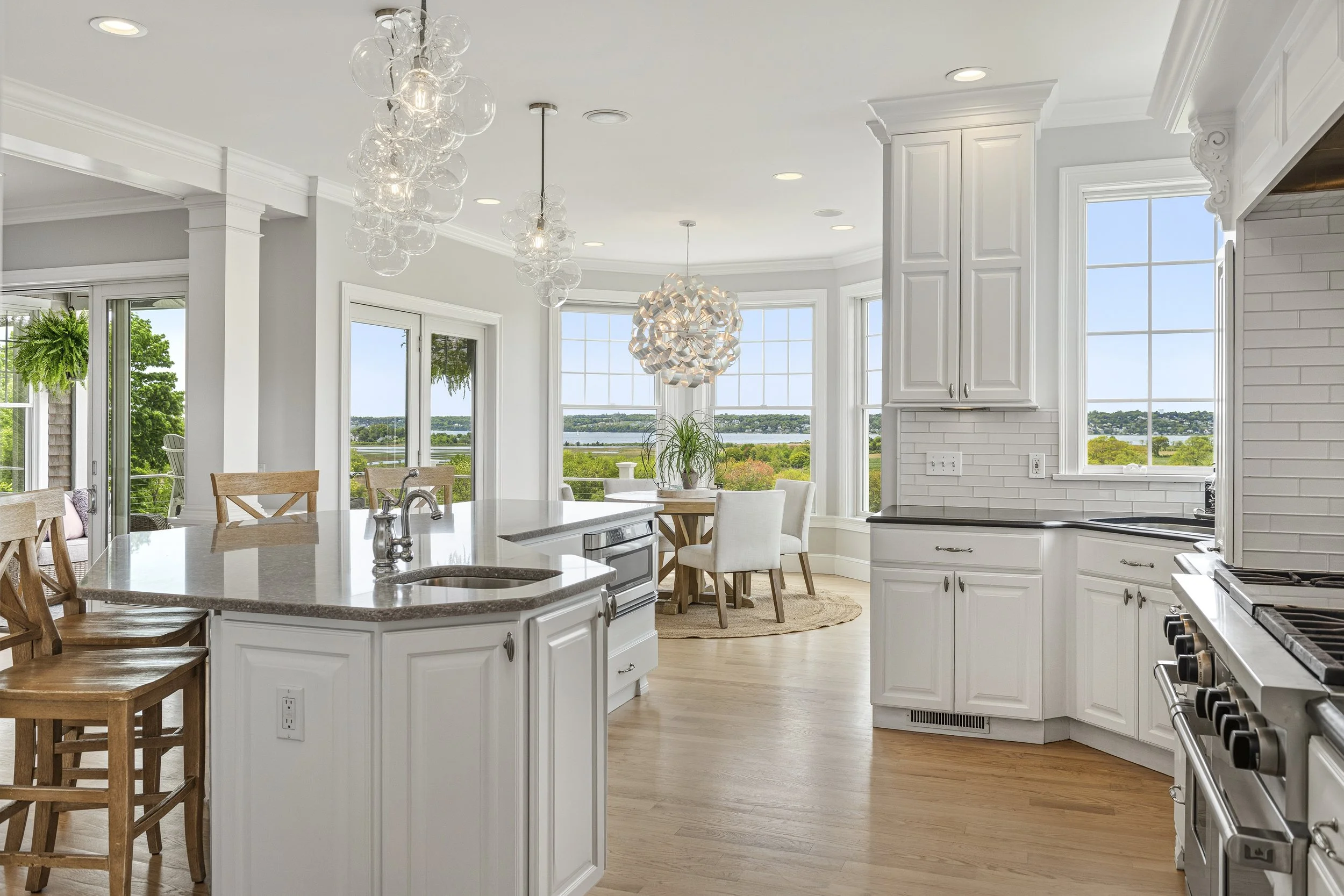 Bright, spacious kitchen with white cabinetry, large windows, and a view of a body of water in the distance. Features include a central island with seating, a modern chandelier, and stainless steel appliances.