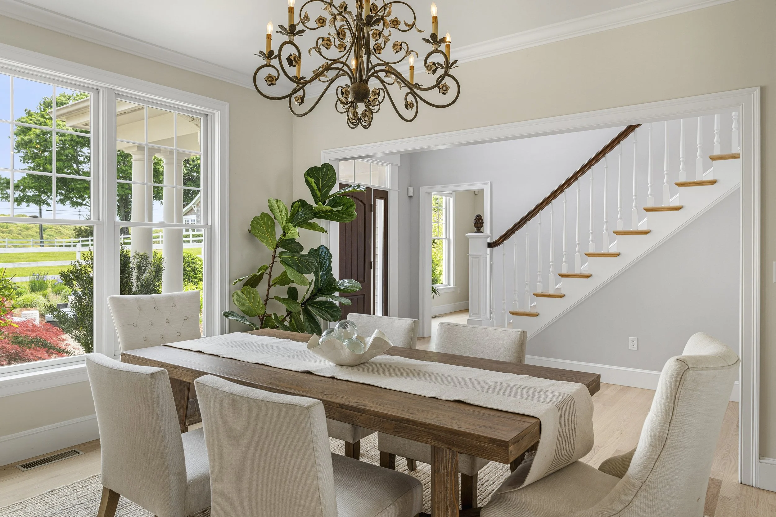 Bright dining room with large windows showing a view of green trees and a white fence outside. There is a wooden dining table surrounded by cream-colored upholstered chairs, and a decorative chandelier hanging from the ceiling. A potted fiddle leaf f