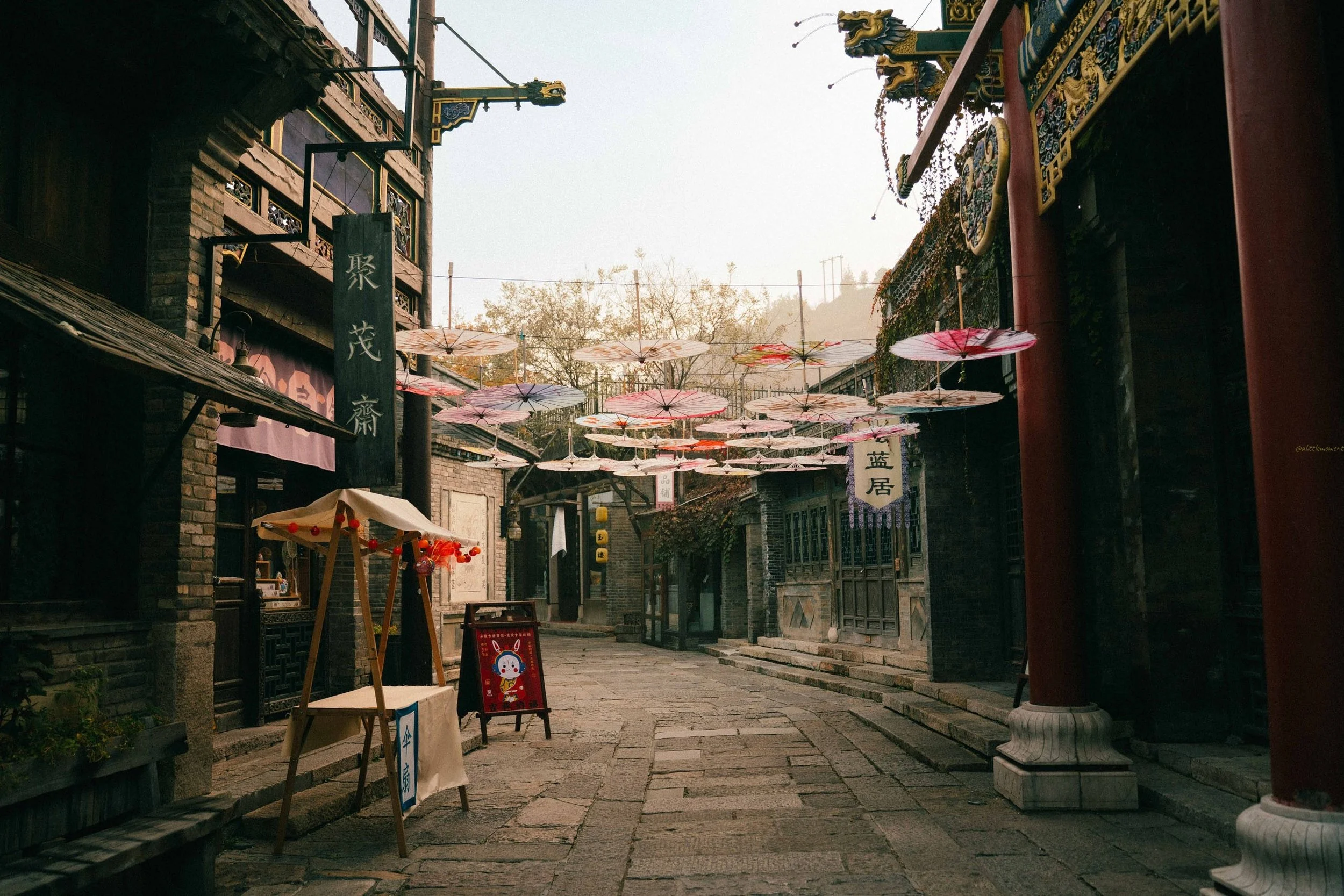 A quiet street scene with traditional Asian architecture, lanterns, and decorative umbrellas hanging overhead, suggesting a festive or cultural area.