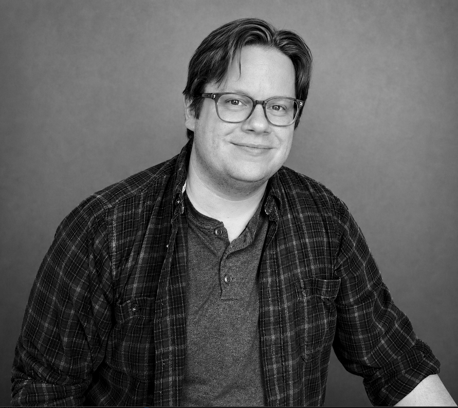 Black and white portrait of a man with glasses, short hair, wearing a plaid shirt over a T-shirt, smiling, with a plain background.