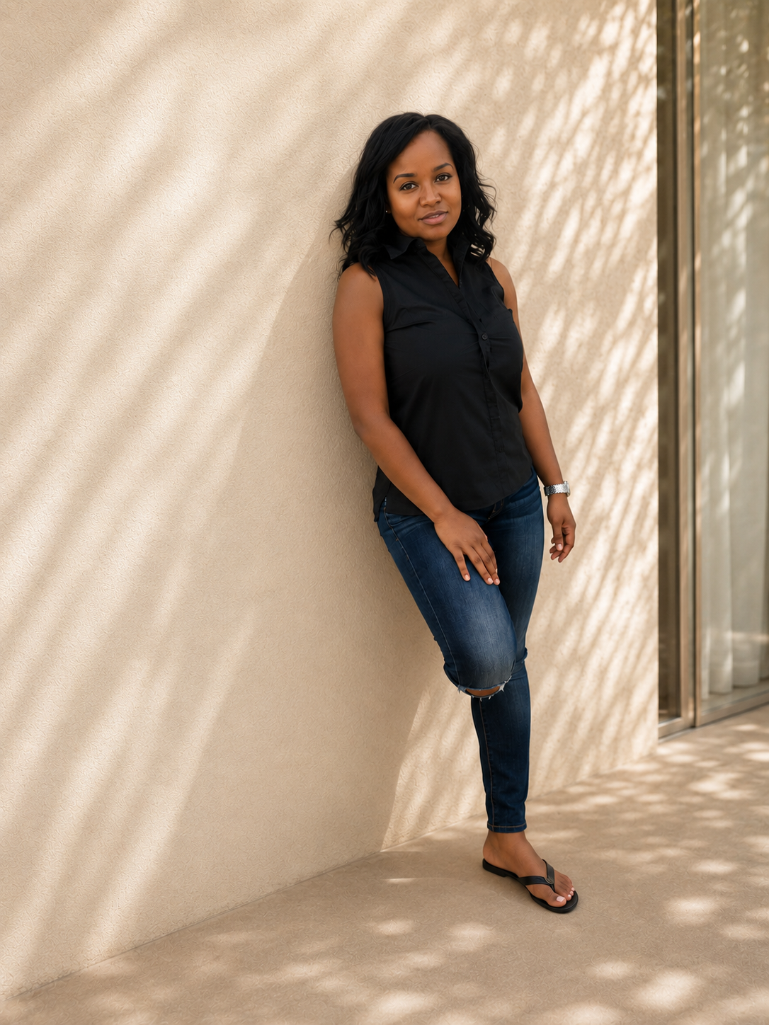 A woman with black wavy hair leaning against a beige wall with patterned shadows, wearing a sleeveless black shirt, ripped blue jeans, and black flip-flops.