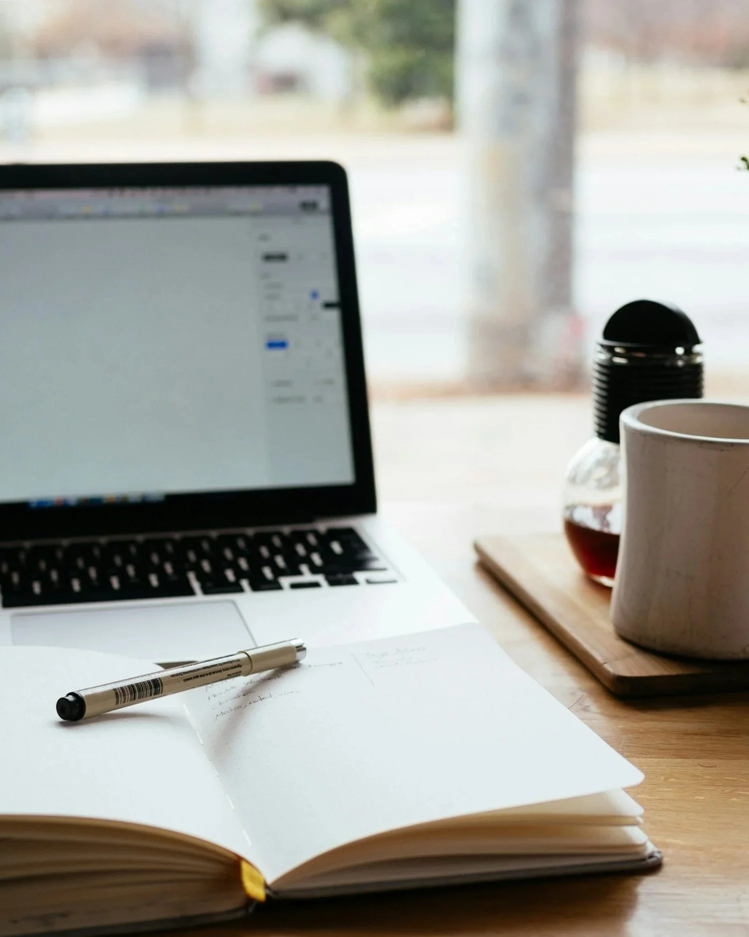 Photo of a laptop displaying a mostly white screen, with a notebook and pen in front of it. A mug and dark-looking liquid also appear at the right side of the frame.
