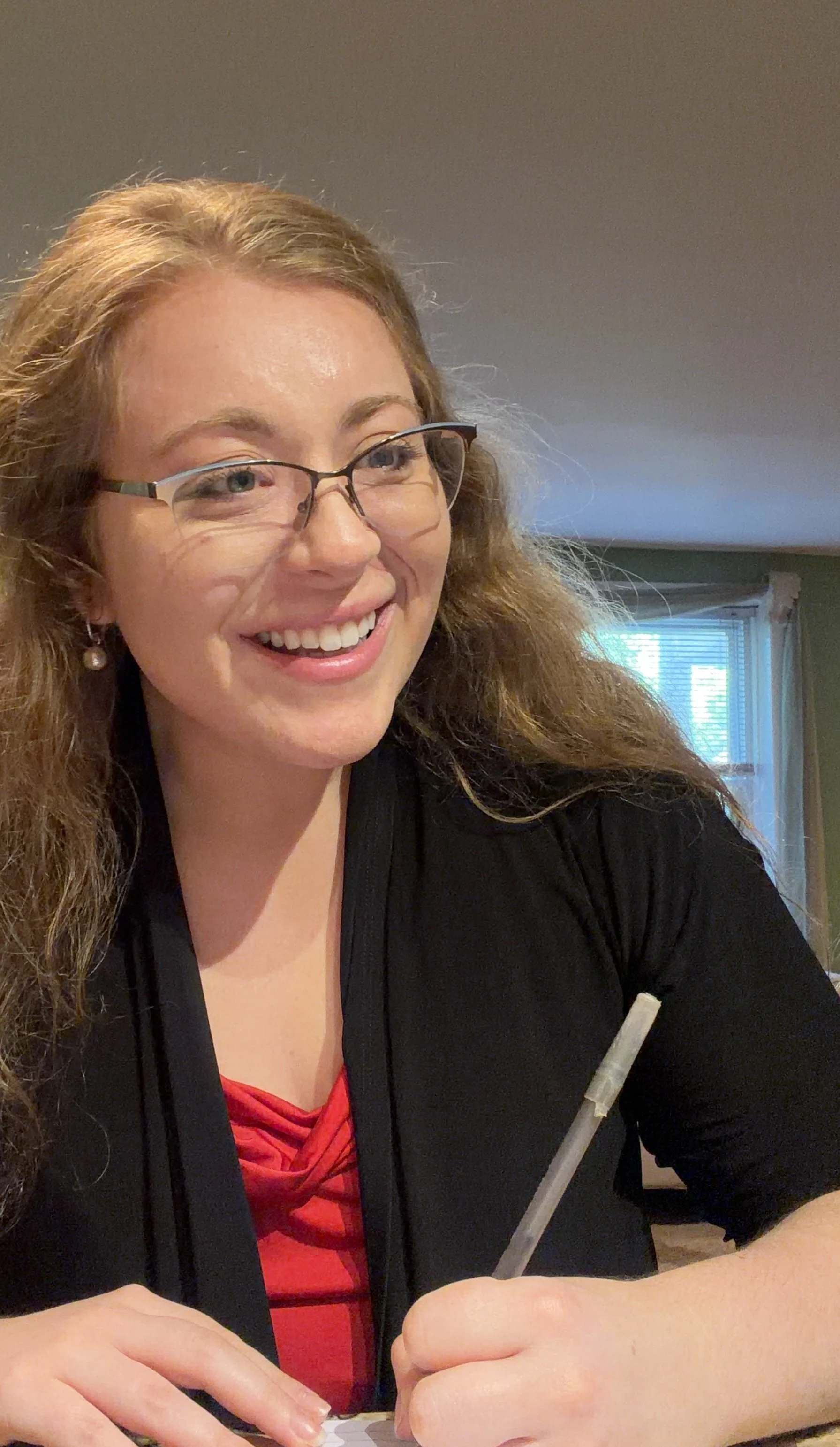 Photo of Sarah Gould, showing her smiling in a friendly way and writing something with the pen in her hand. She wears a red shirt with a black shrug, glasses, and pearl earrings.