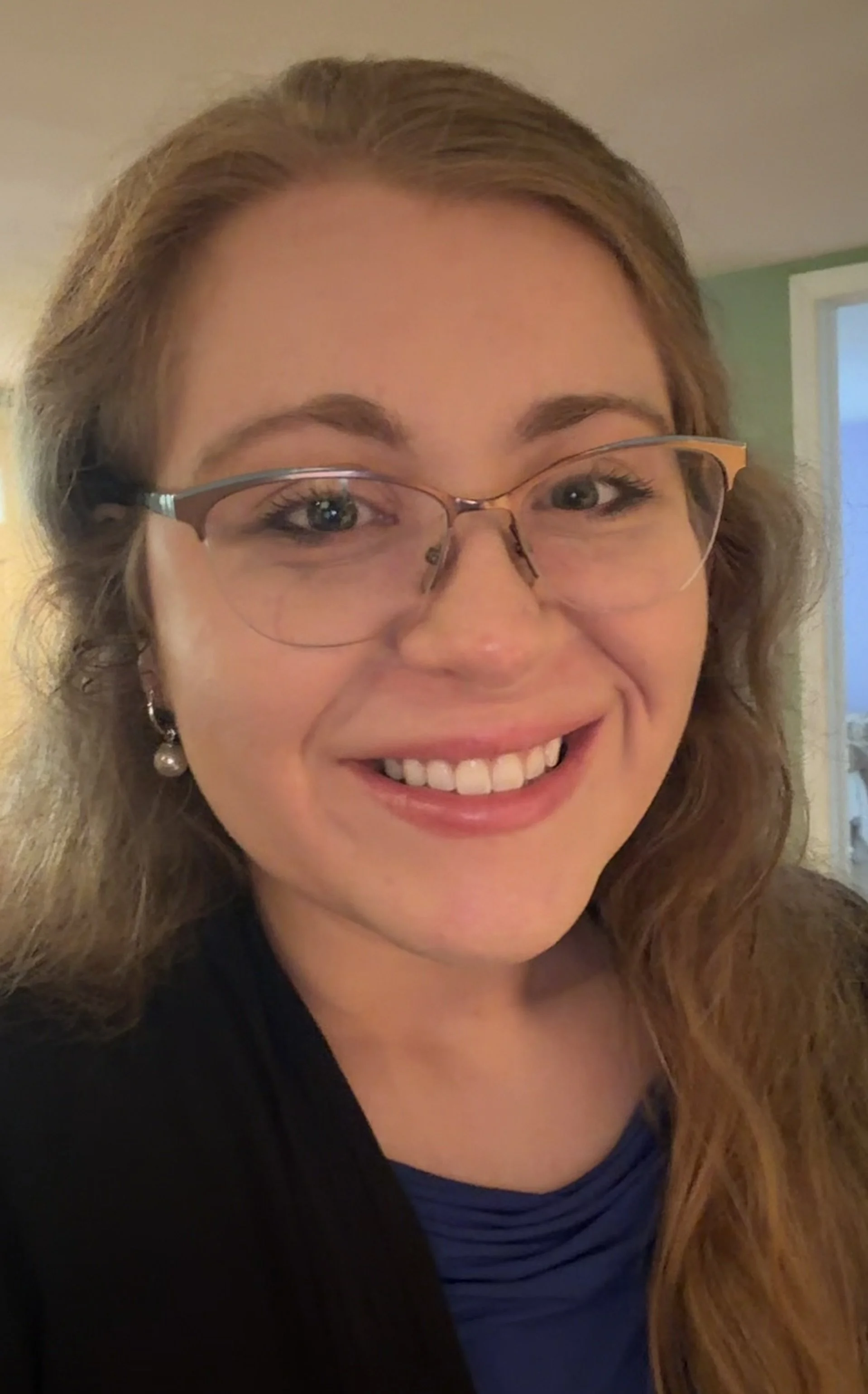A headshot of Sarah Gould smiling in an approachable way. She has long blonde hair and wears a blue shirt with a black shrug over it. She also wears glasses and pearl earrings, projecting a professional, competent image.