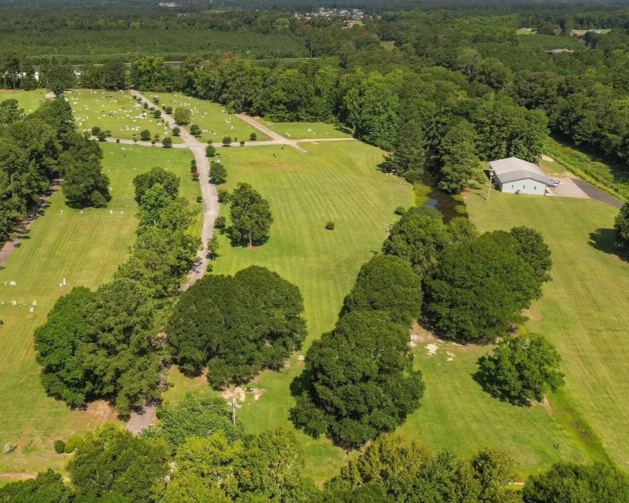Aerial view of a green cemetery with rows of white grave markers; surrounded by trees and a small pond with a building nearby.