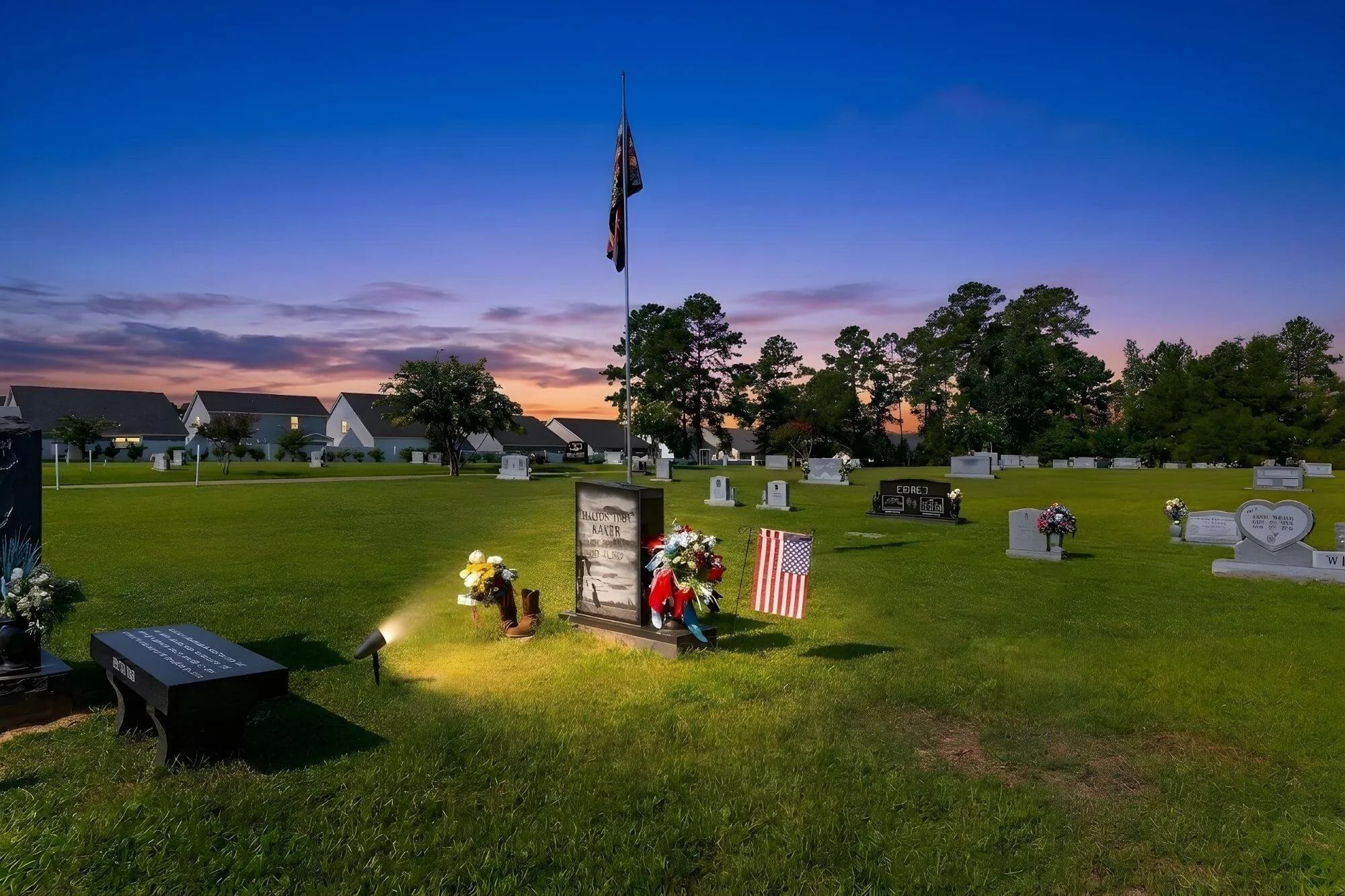 A cemetery at dusk with headstones and flowers, a flagpole with a flag, trees in the background, and a colorful sunset sky.