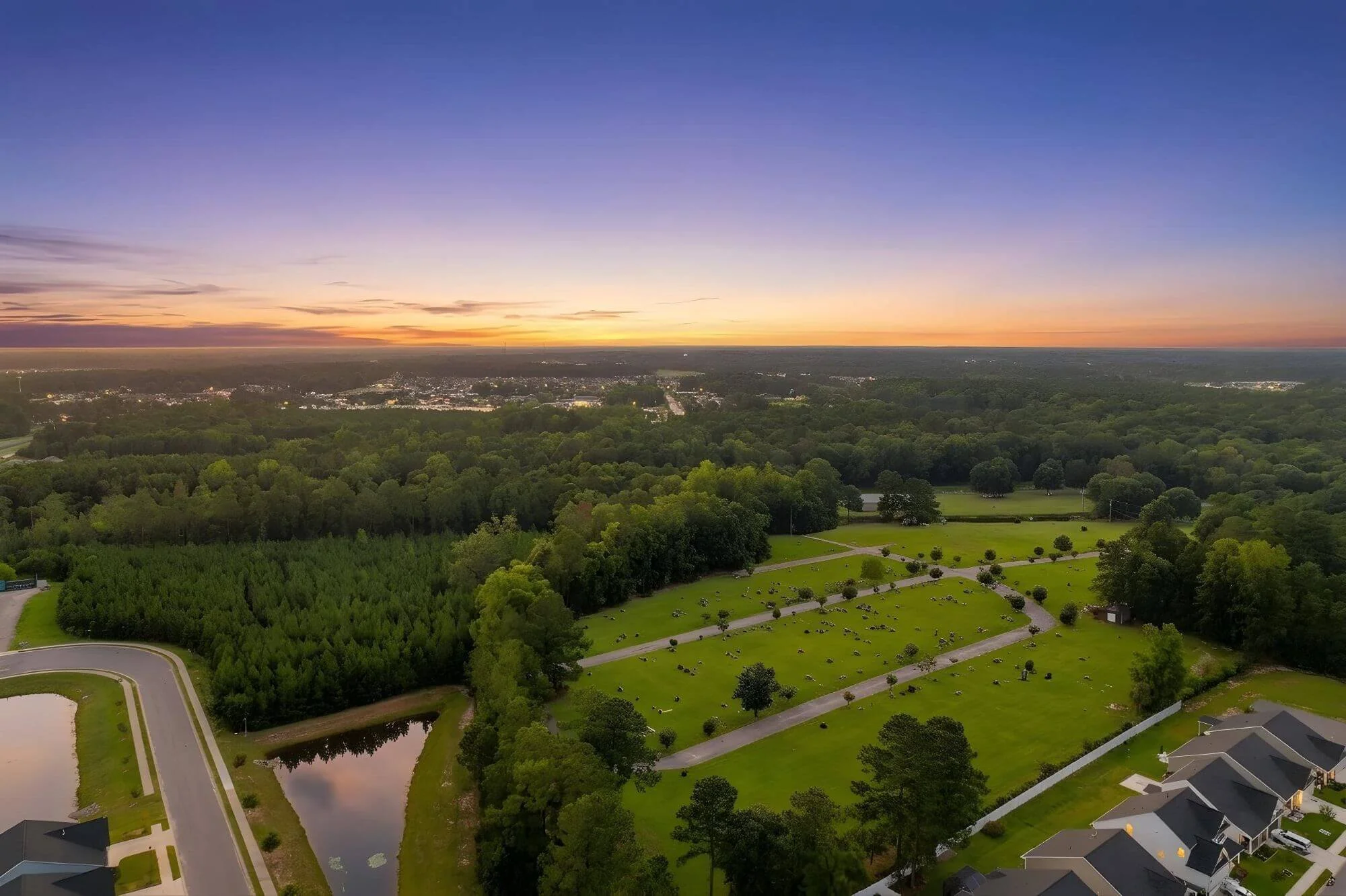 Sunset over Westside Memorial Gardens in Conway with green yards and trees, a small pond, and houses with gray roofs.