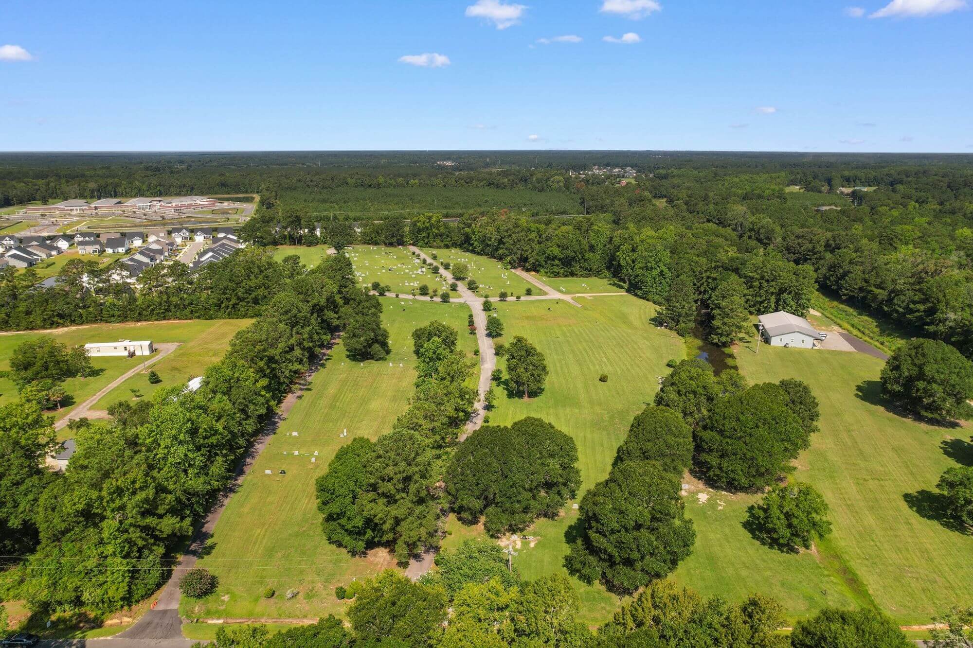 Aerial view of a green landscape with a cemetery, residential houses, a wooded forest, and a clear blue sky with scattered clouds.