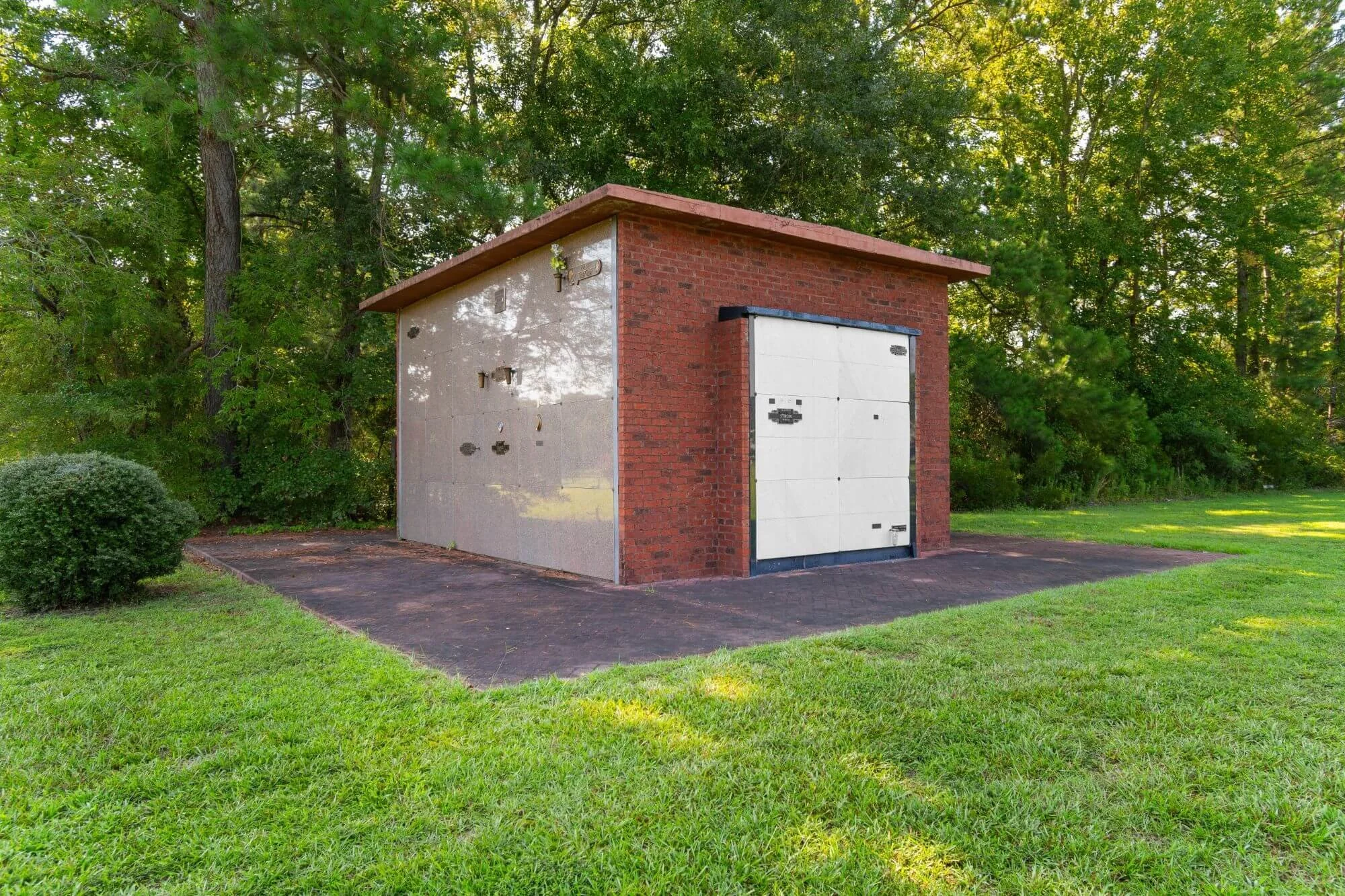 A brick and concrete mausoleum structure with a closed white garage door, surrounded by green grass and trees in the background.