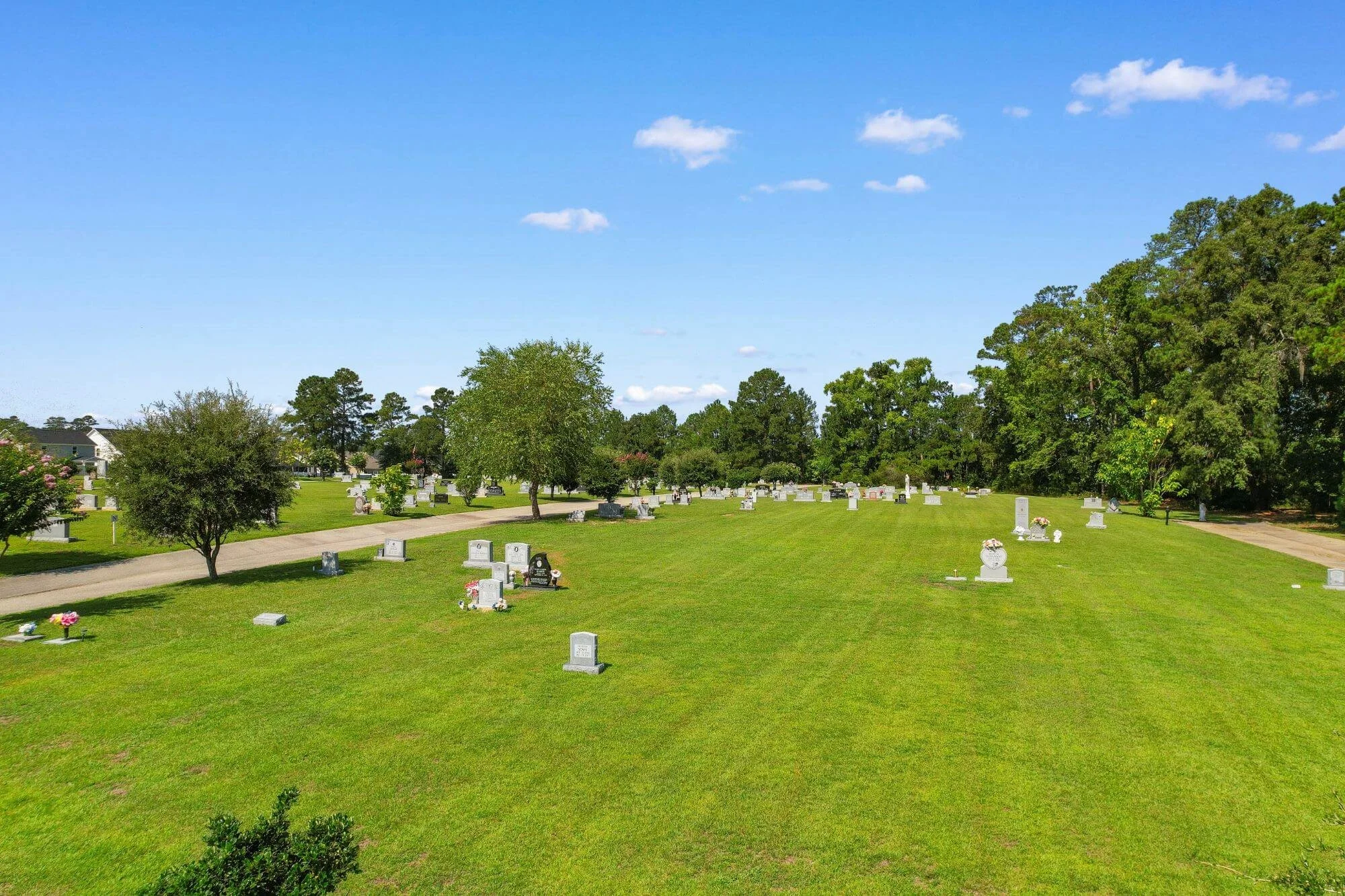 A cemetery with rows of flat white headstones on green grass, surrounded by tall green trees under a bright blue sky with a few scattered clouds.