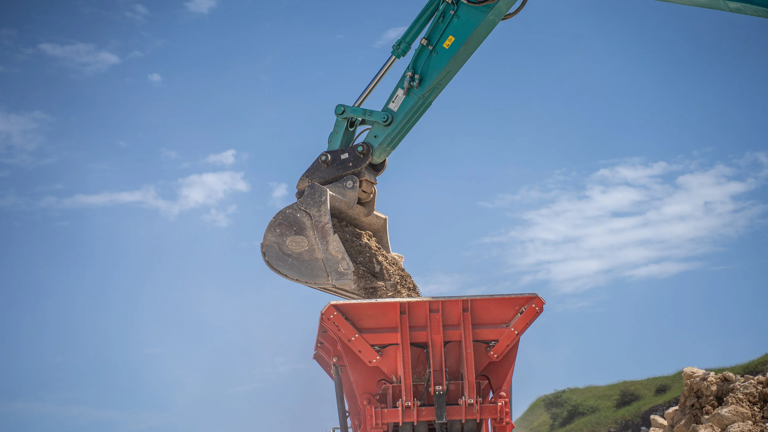A construction excavator loading dirt into a red dump truck against a bright blue sky with some clouds.