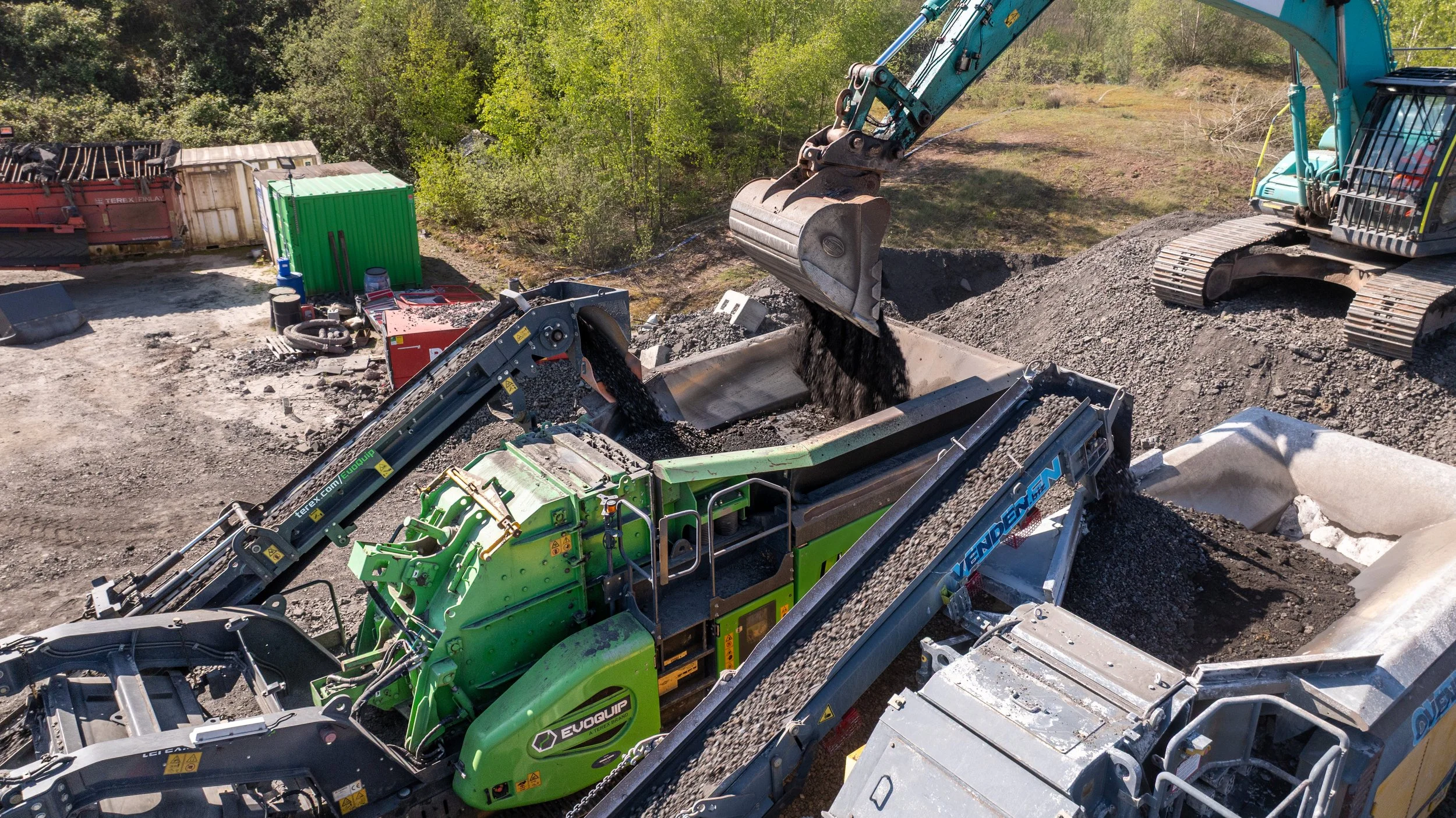 Construction site with excavator and machinery, excavating soil surrounded by trees and containers.