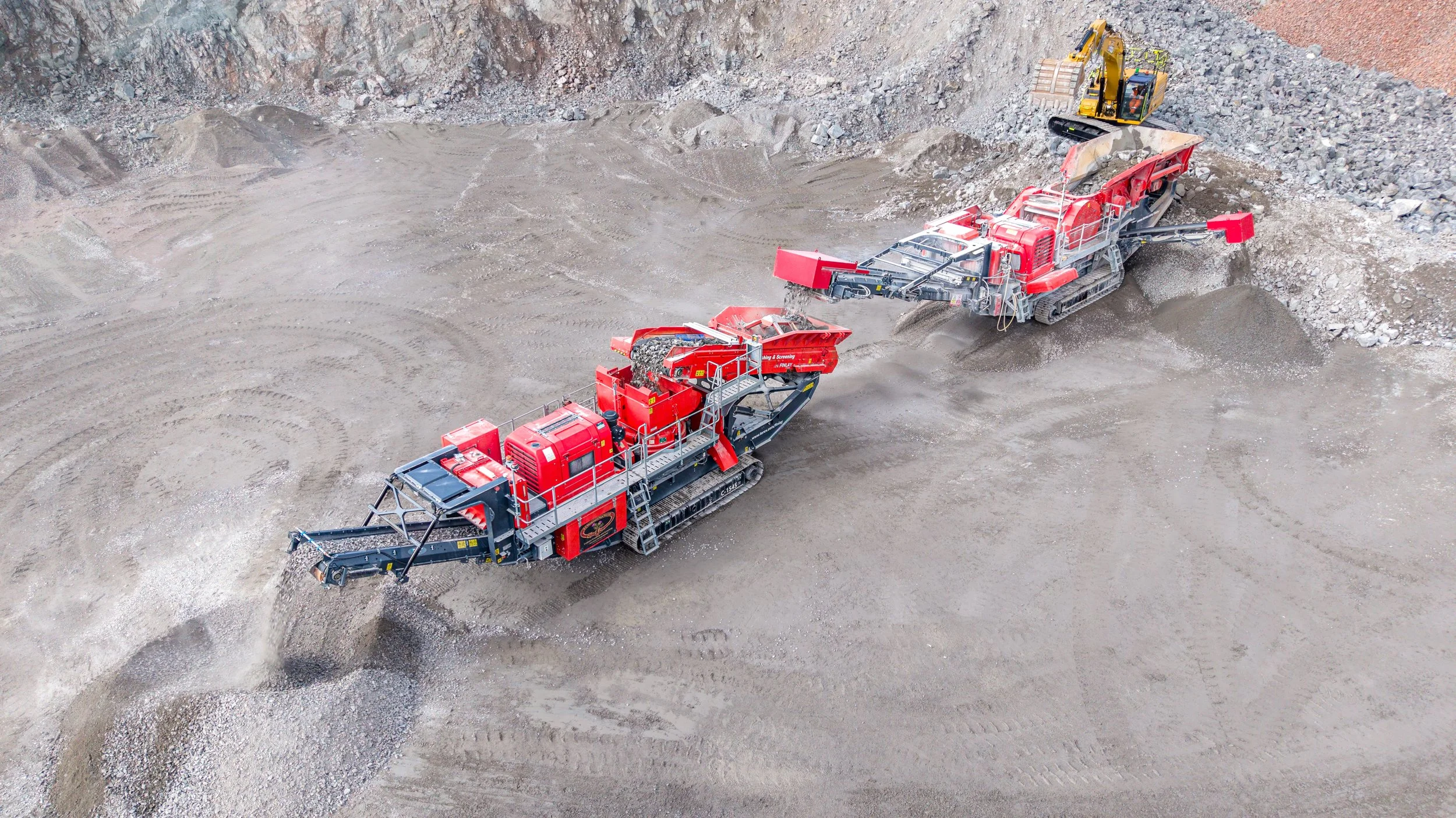 Two large red and black industrial machines working on a dirt construction site, excavating and processing gravel or soil.