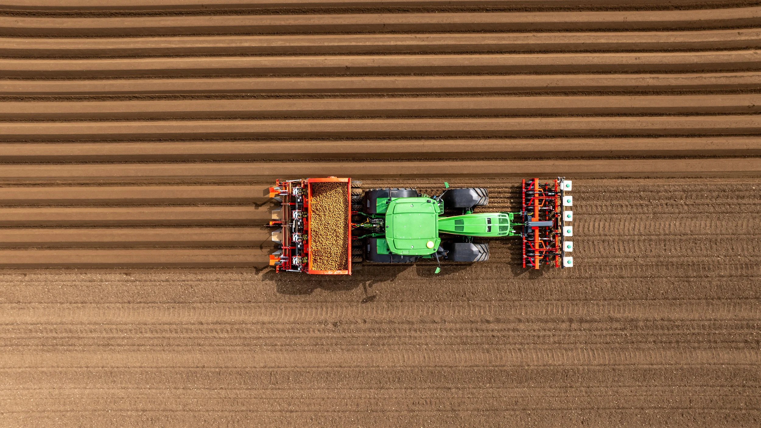 An aerial view of a green tractor working on a field with evenly spaced rows of soil, planting or preparing the land.
