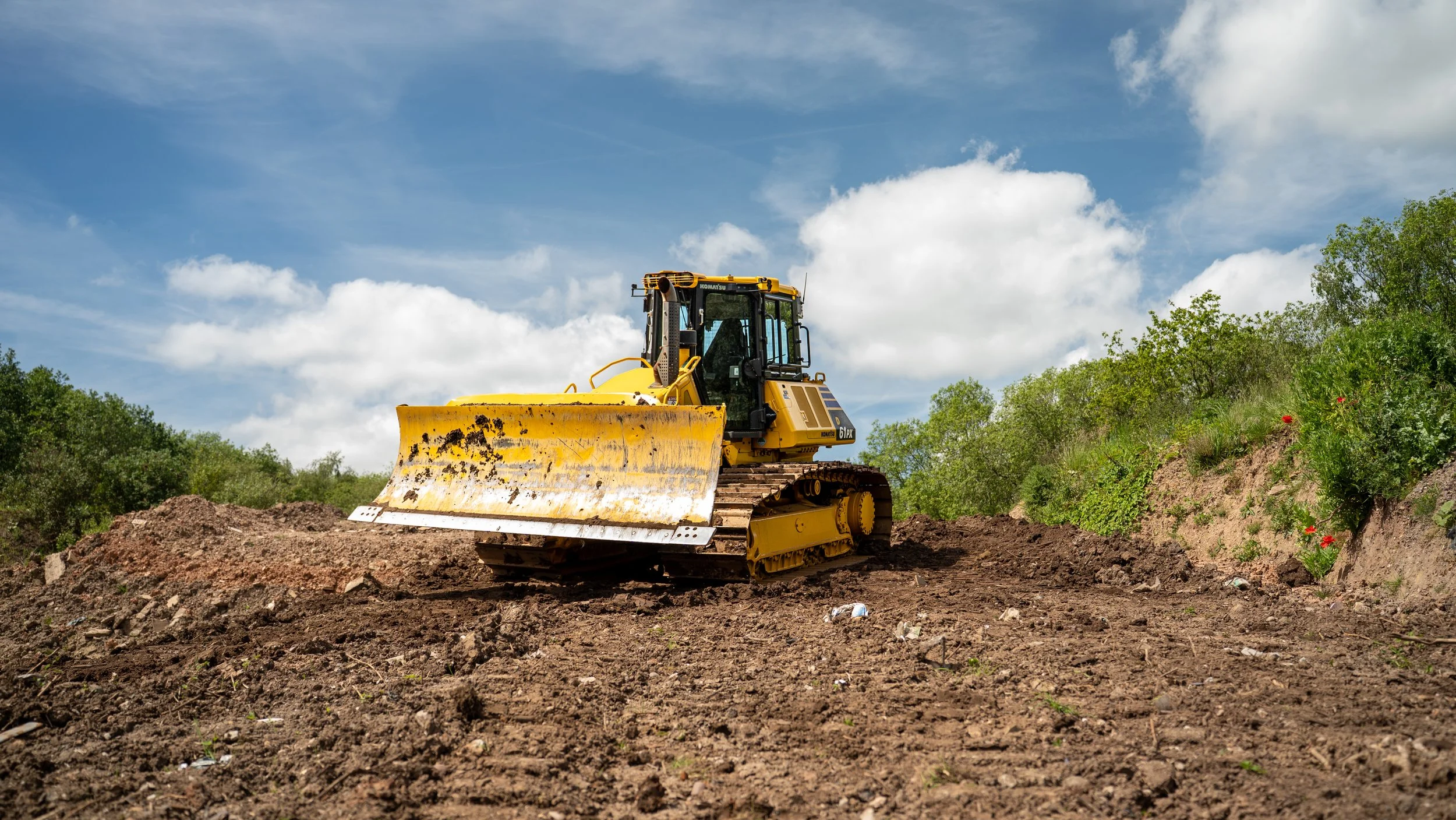 Yellow bulldozer moving dirt on a construction site with green trees and blue sky in the background.
