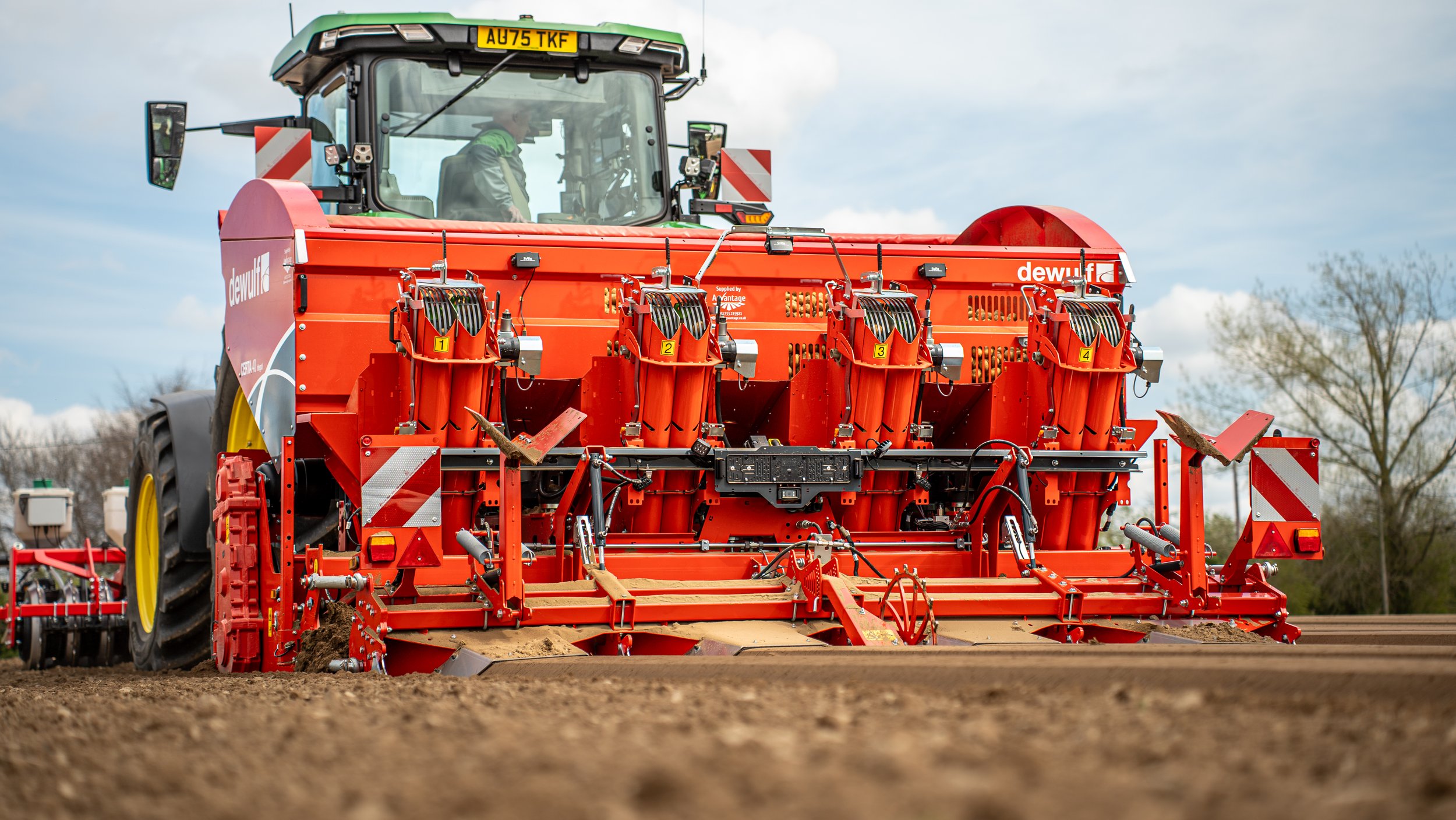 A large red agricultural machine operating in a field, with a person inside the cab. The machine has multiple components labeled with numbers, and appears to be used for soil preparation or planting.