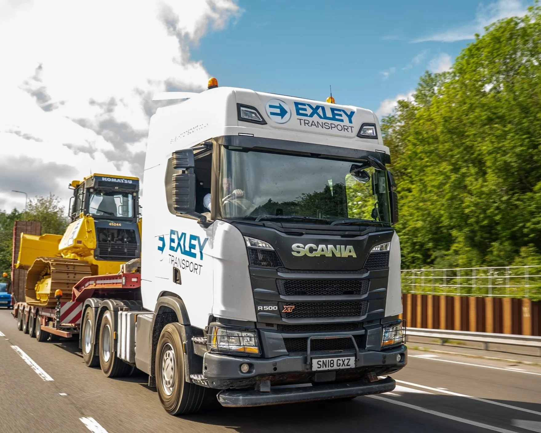 A large white semi-truck with EXLEY TRANSPORT branding on the front and side, transporting construction equipment on a highway with green trees and a partly cloudy sky in the background.