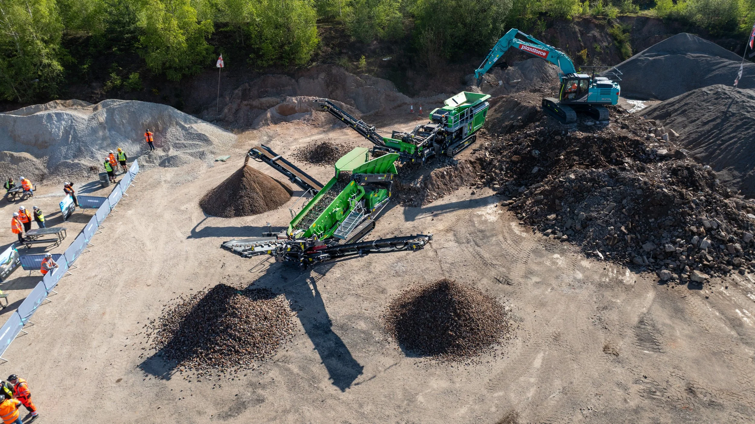 Construction site with large machinery and workers, involving earth and rock excavation, surrounded by trees.
