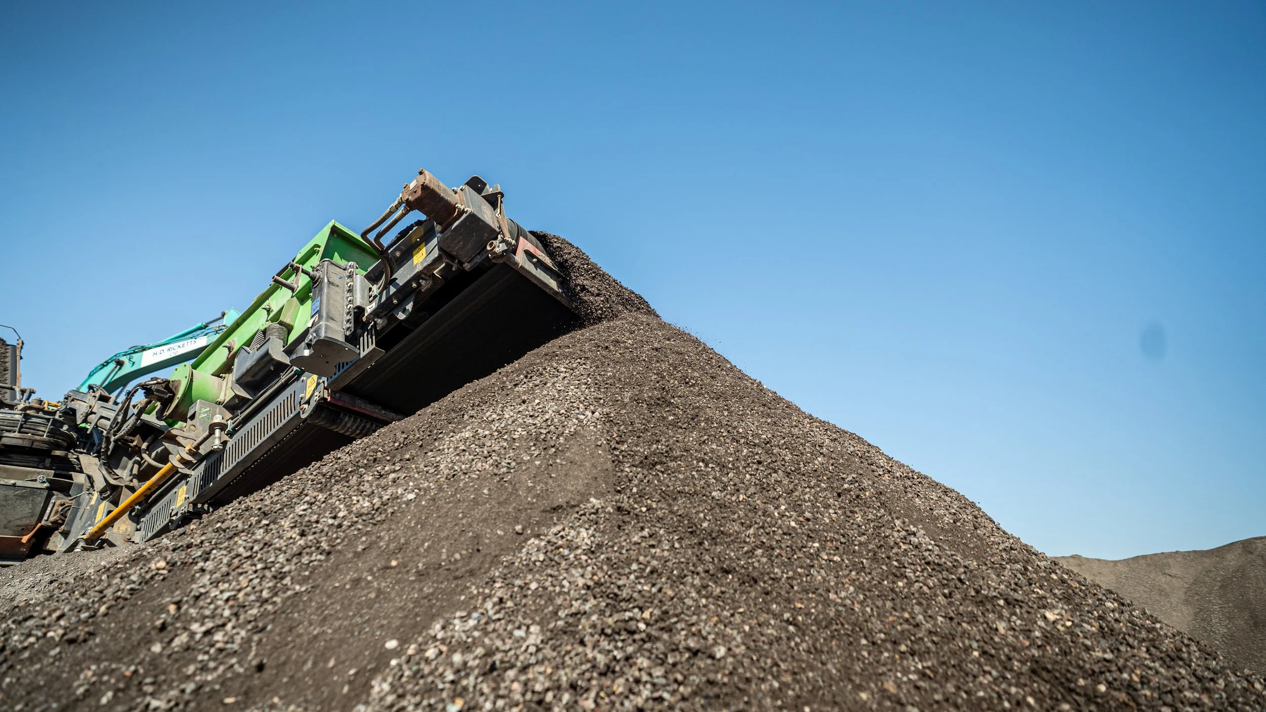 A large industrial machine spreading gravel or small stones on the ground under a clear blue sky.