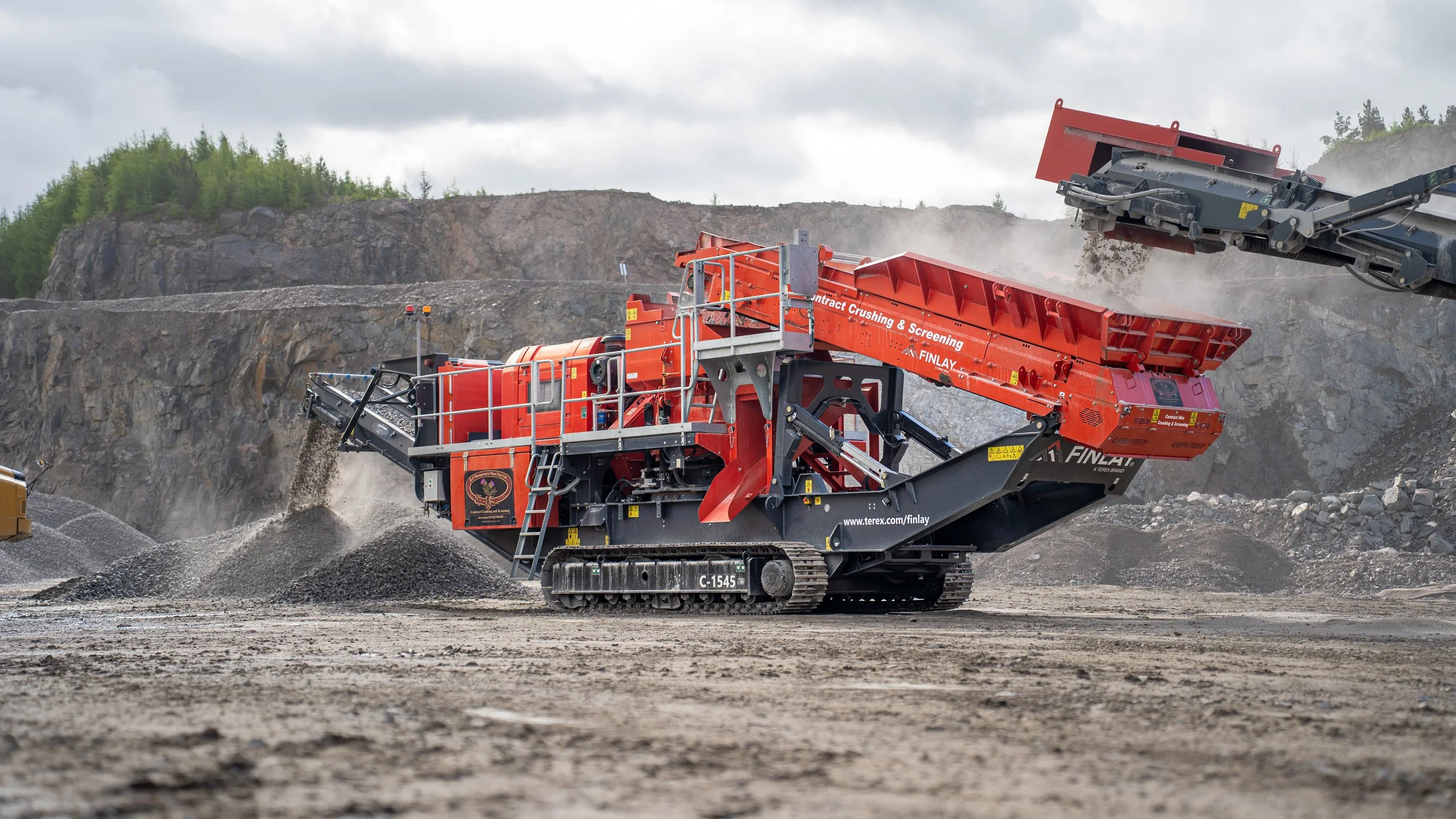 Large orange mobile crusher working in a quarry, crushing rocks and gravel with dust in the air, mountains and cloudy sky in the background.
